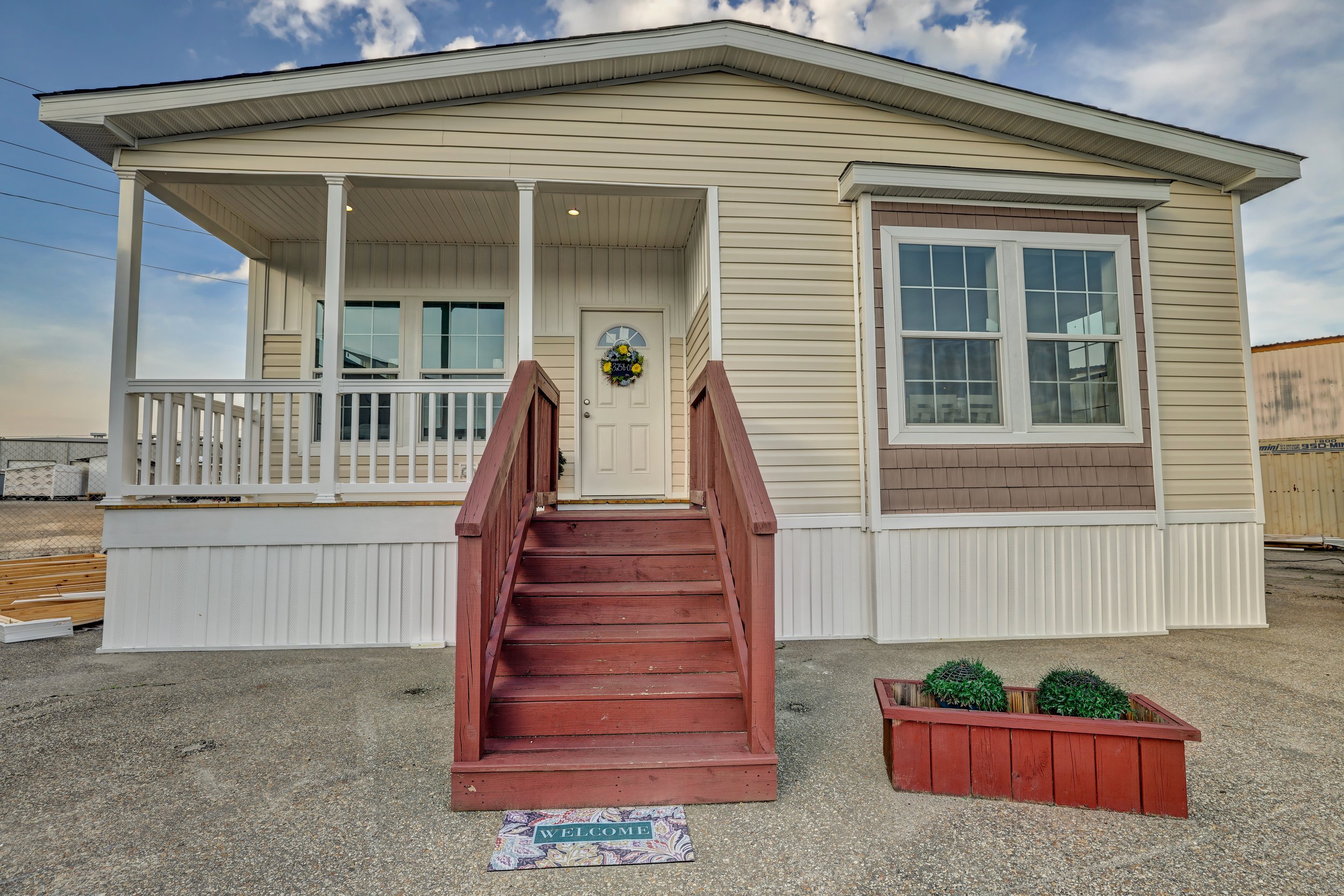Exterior view of a beige manufactured home with a covered porch and red wooden steps. A welcome mat lies at the base of the steps. Two large windows are visible, along with a flower wreath on the front door. A red planter box with bushes sits to the
