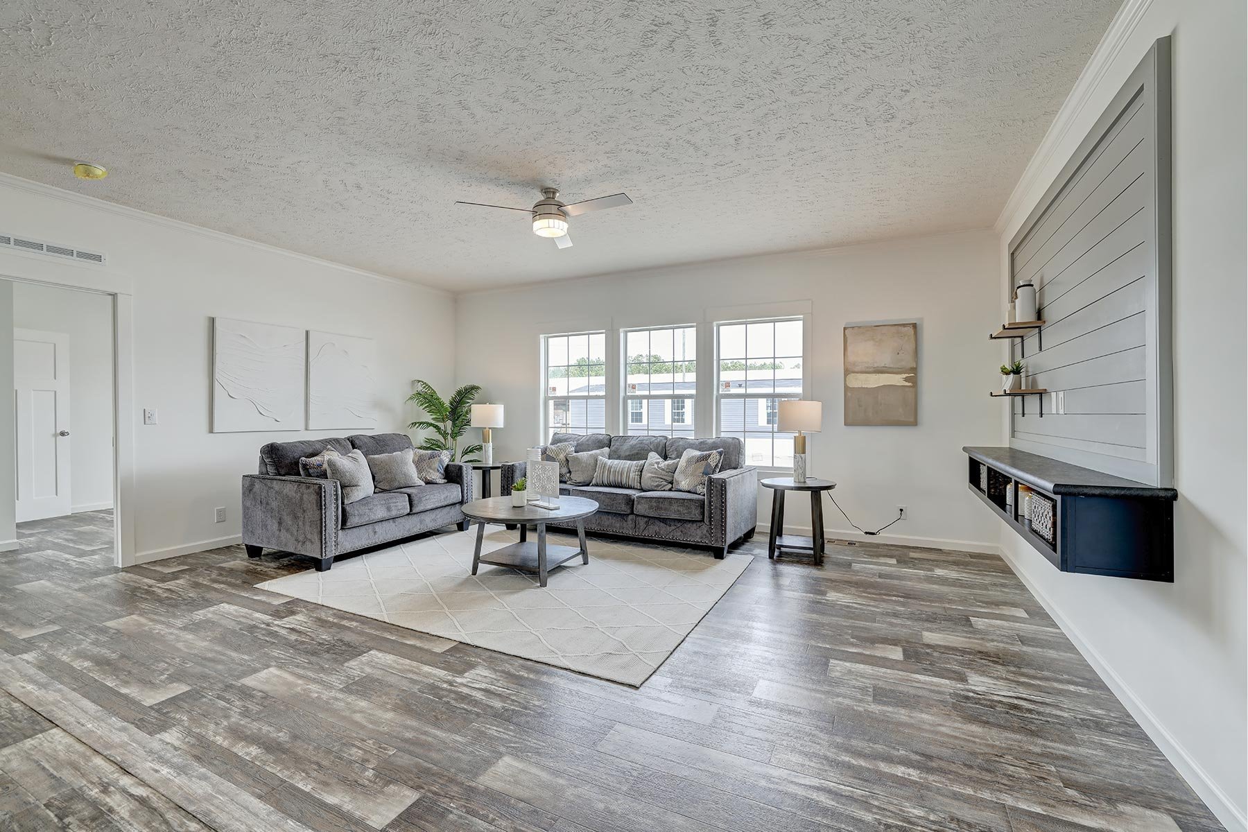 Modern living room with gray sofas, coffee table, and a decorative rug. Ceiling fan and windows in background. Neutral decor with plants and art on walls. Built-in shelves on right wall.