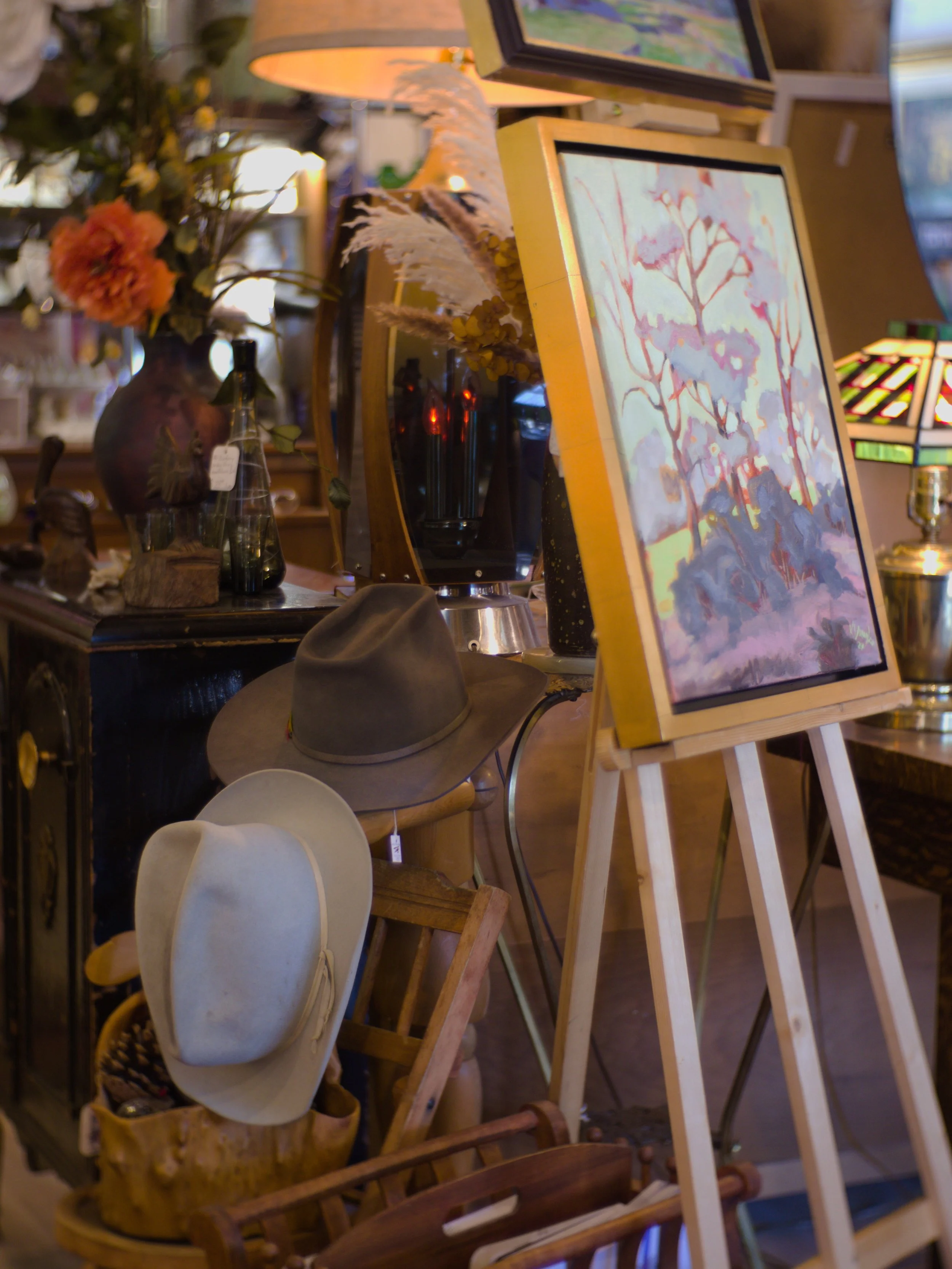 A display of various decorative items including a floral arrangement in a large vase, several bottles, a gray hat, and framed artwork on an easel inside a store or gallery.