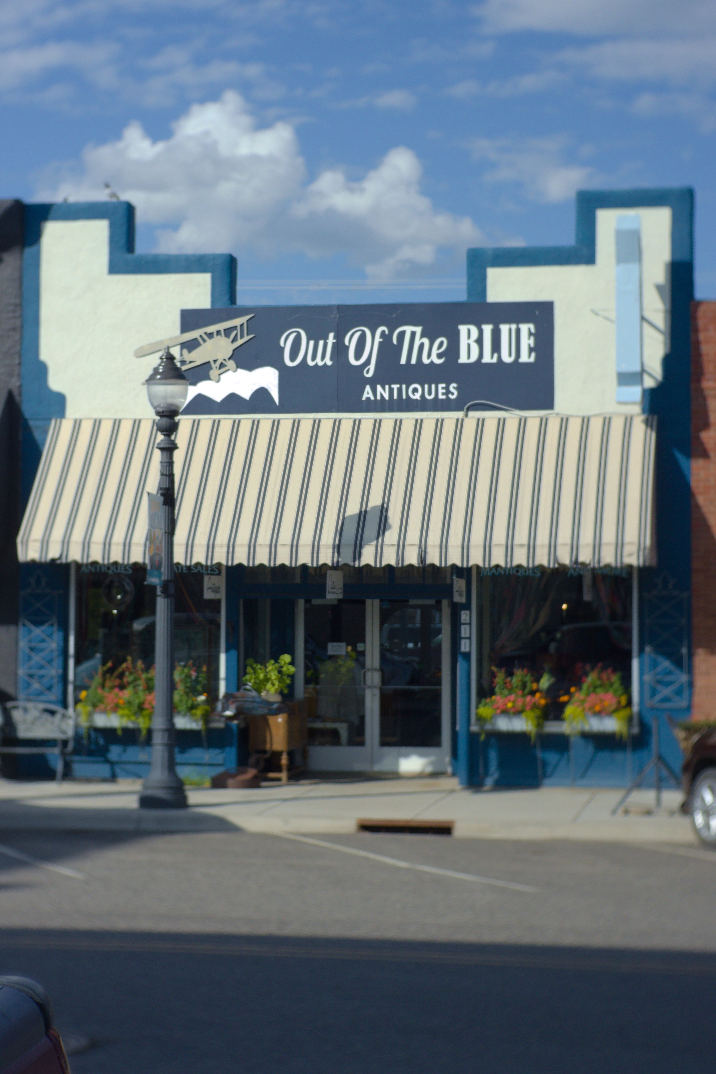 Storefront with a sign that reads 'Out Of The Blue Antiques' and a vintage airplane decor, with striped awning and flower boxes outside, under a partly cloudy sky.