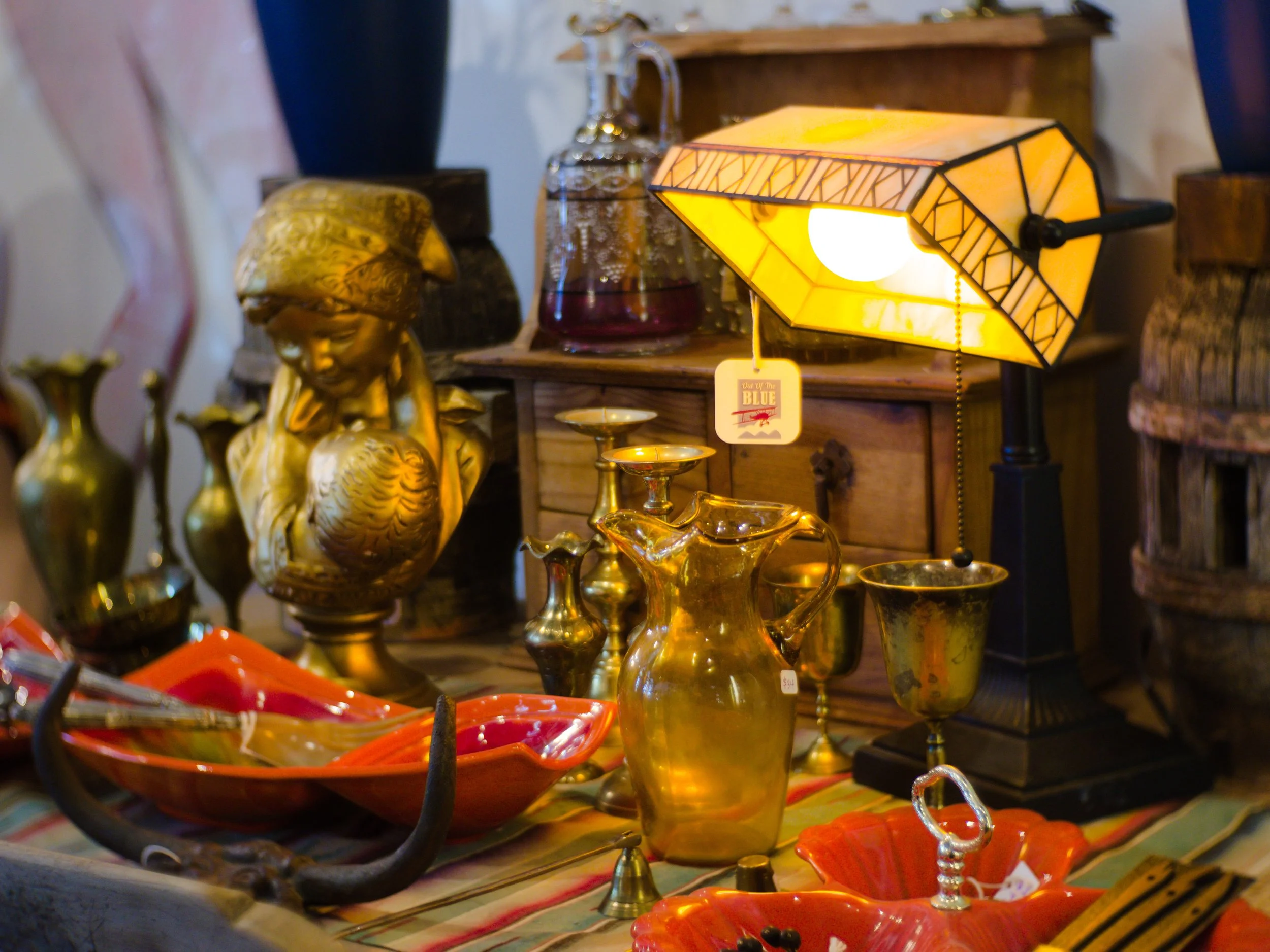 Various decorative objects including a yellow glass lamp, gold-colored figurines, vases, bowls, and other glassware displayed on a table at an antique shop.