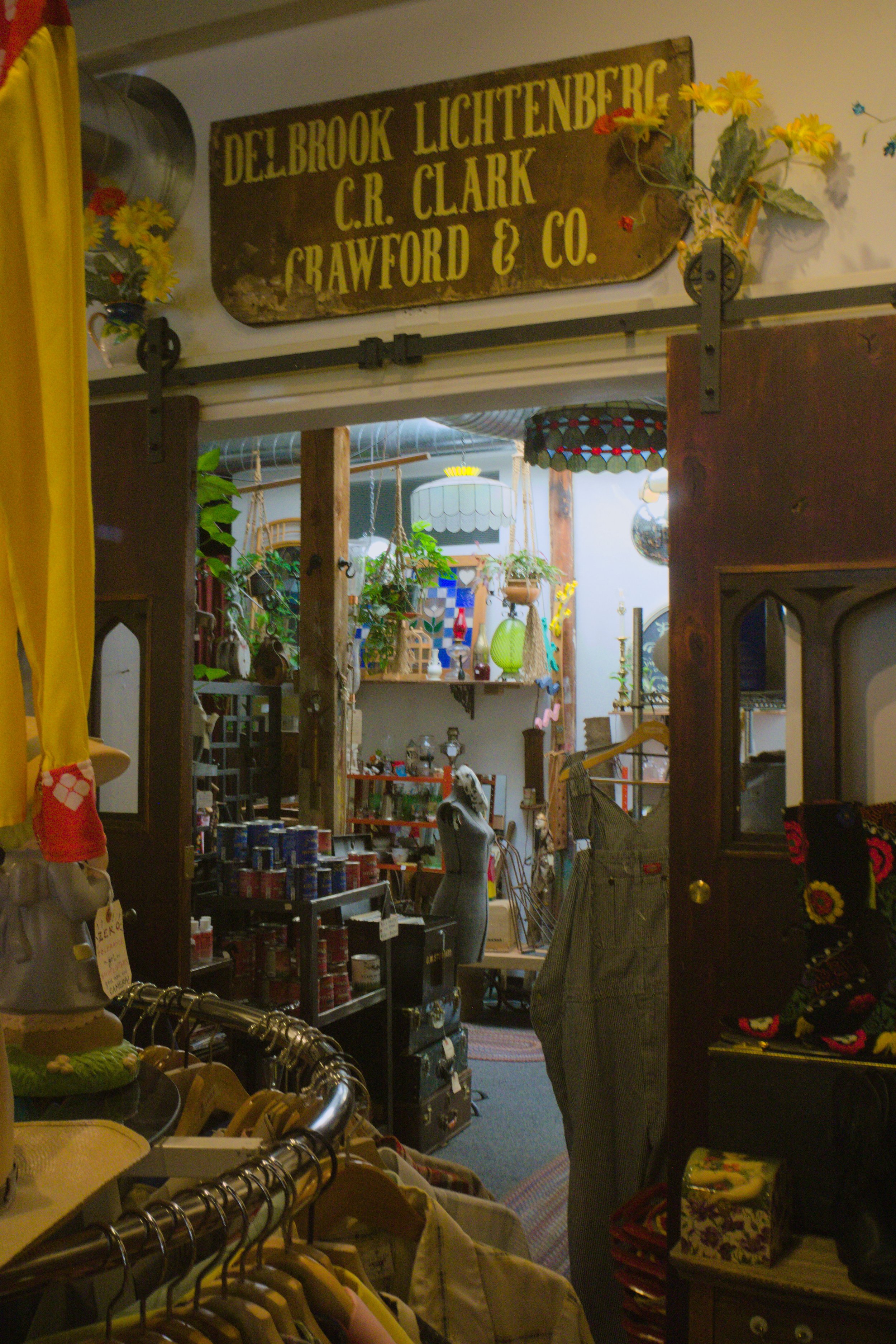 View of an indoor thrift store or antique shop with a wooden sign hanging on the wall, decorated with artificial flowers. The store has clothing racks, shelves filled with various items, and decorative lamps and plants, creating a vintage and cozy at