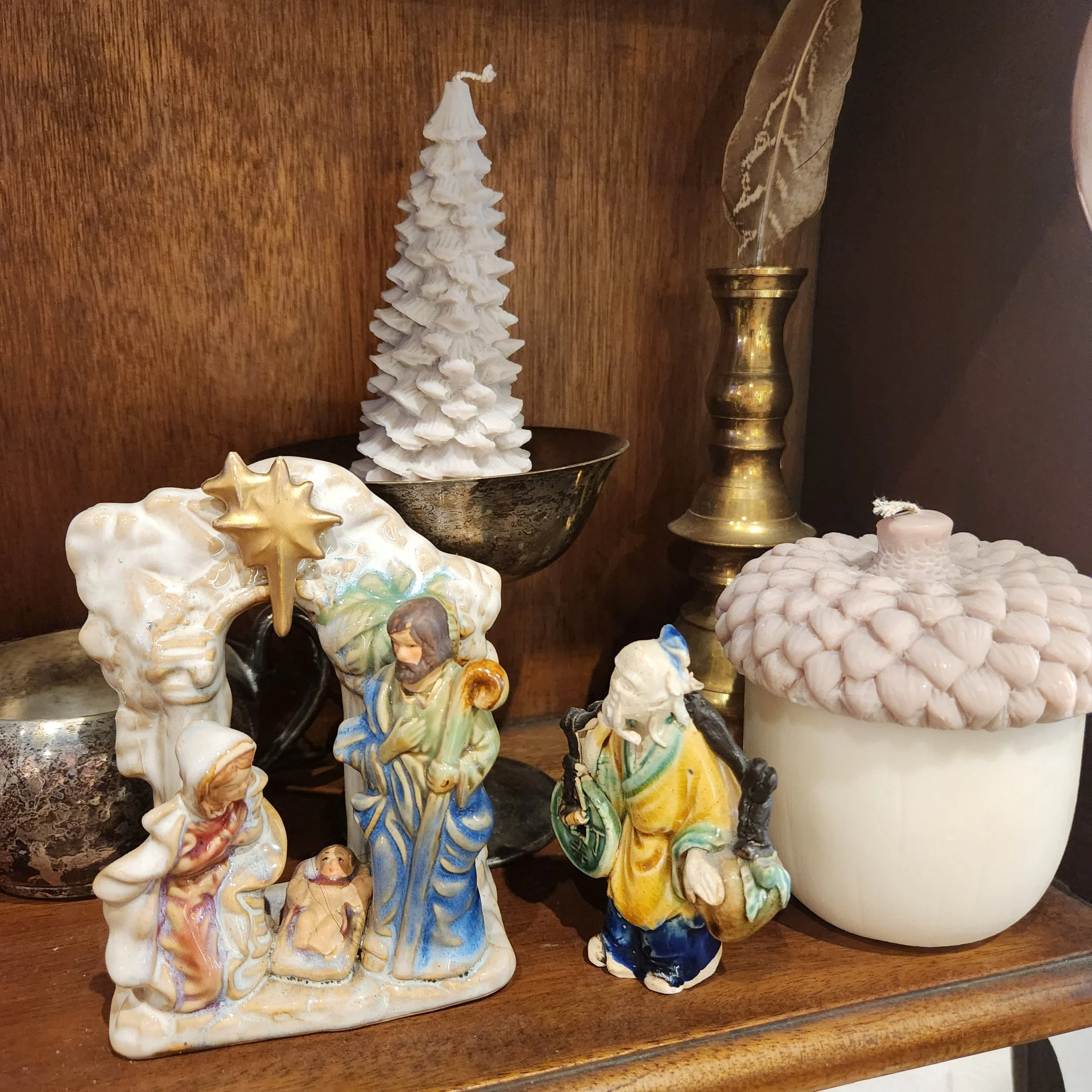 Collection of decorative ceramic figurines on a wooden surface, including a tall, white, layered Christmas tree, a small figurine of a woman in a blue dress, and other assorted ornaments.