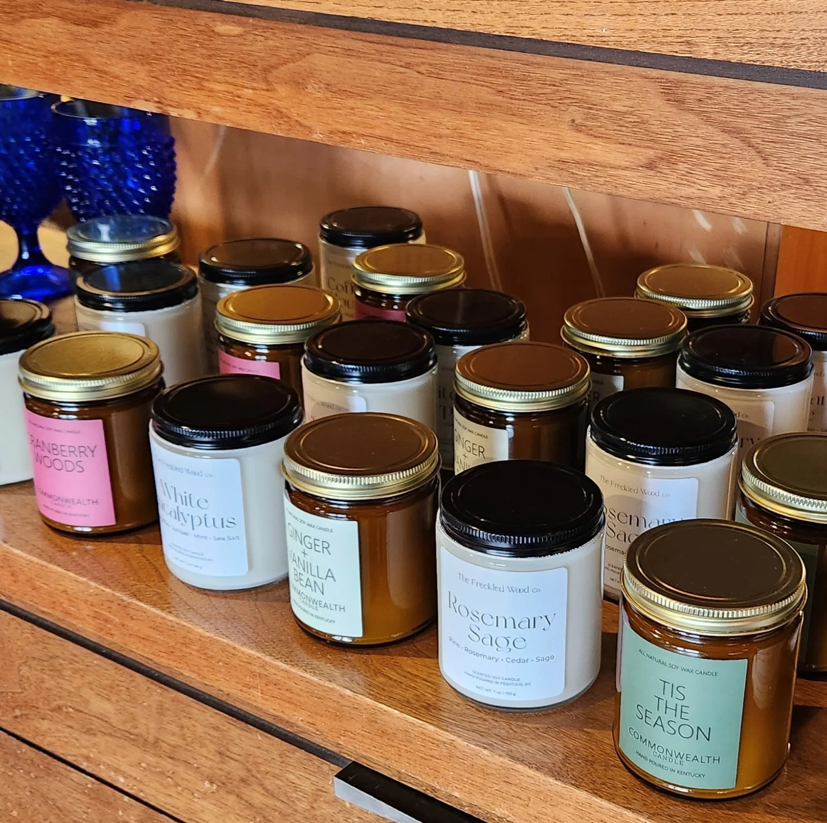 A collection of scented candles in glass jars with black, gold, and pink lids arranged on a wooden shelf. The candles have labels with various names like 'Rosemary Sage,' 'Ginger Vanilla Bean,' and 'Tis the Season.' The shelf has a natural wood finis