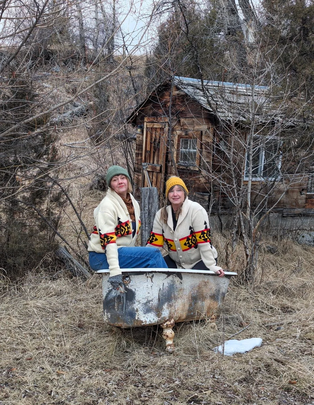 Two women in warm sweaters and hats sitting on an old rusty bathtub outdoors, with a small, weathered wooden house in a wooded area behind them.