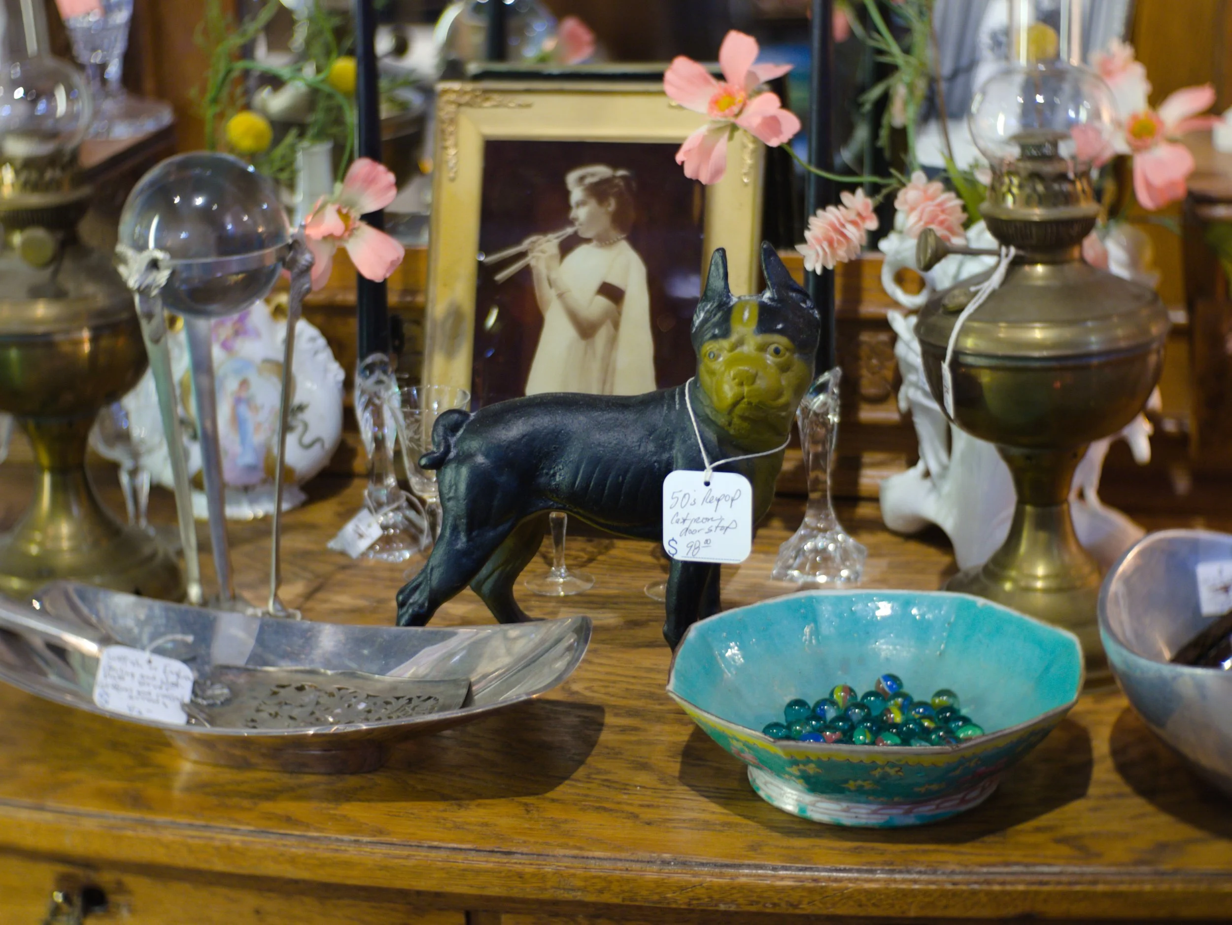 A collection of vintage and decorative items including a black dog figurine, pink artificial flowers, glass candle holders, various bowls, and an framed black-and-white photo of a woman playing a flute, displayed on a wooden table.