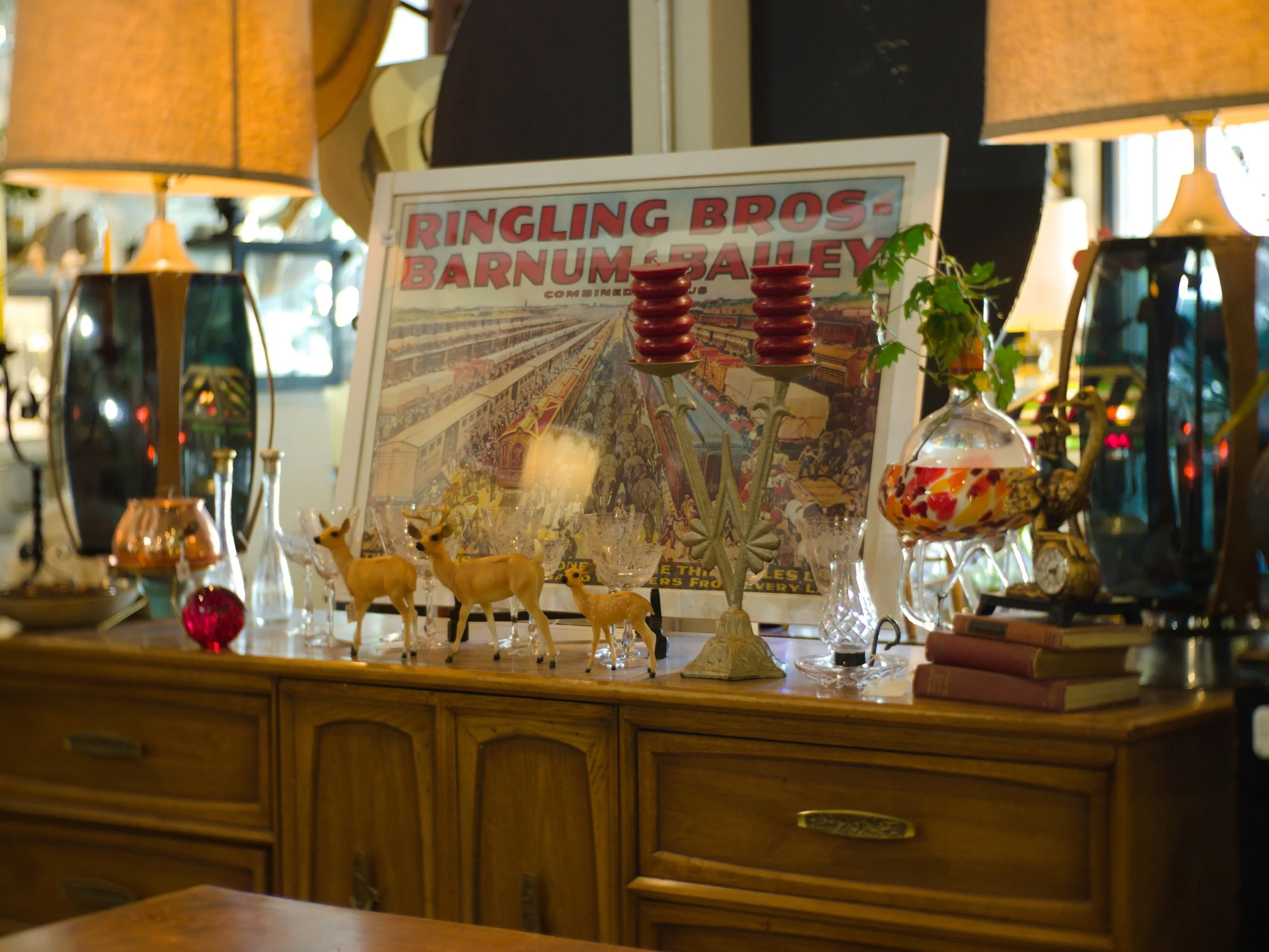 A wooden display cabinet with various decorative items including miniature deer figurines, glassware, lamps, and a framed vintage poster for Ringling Bros. Barnum & Bailey Circus, with lamps on either side.