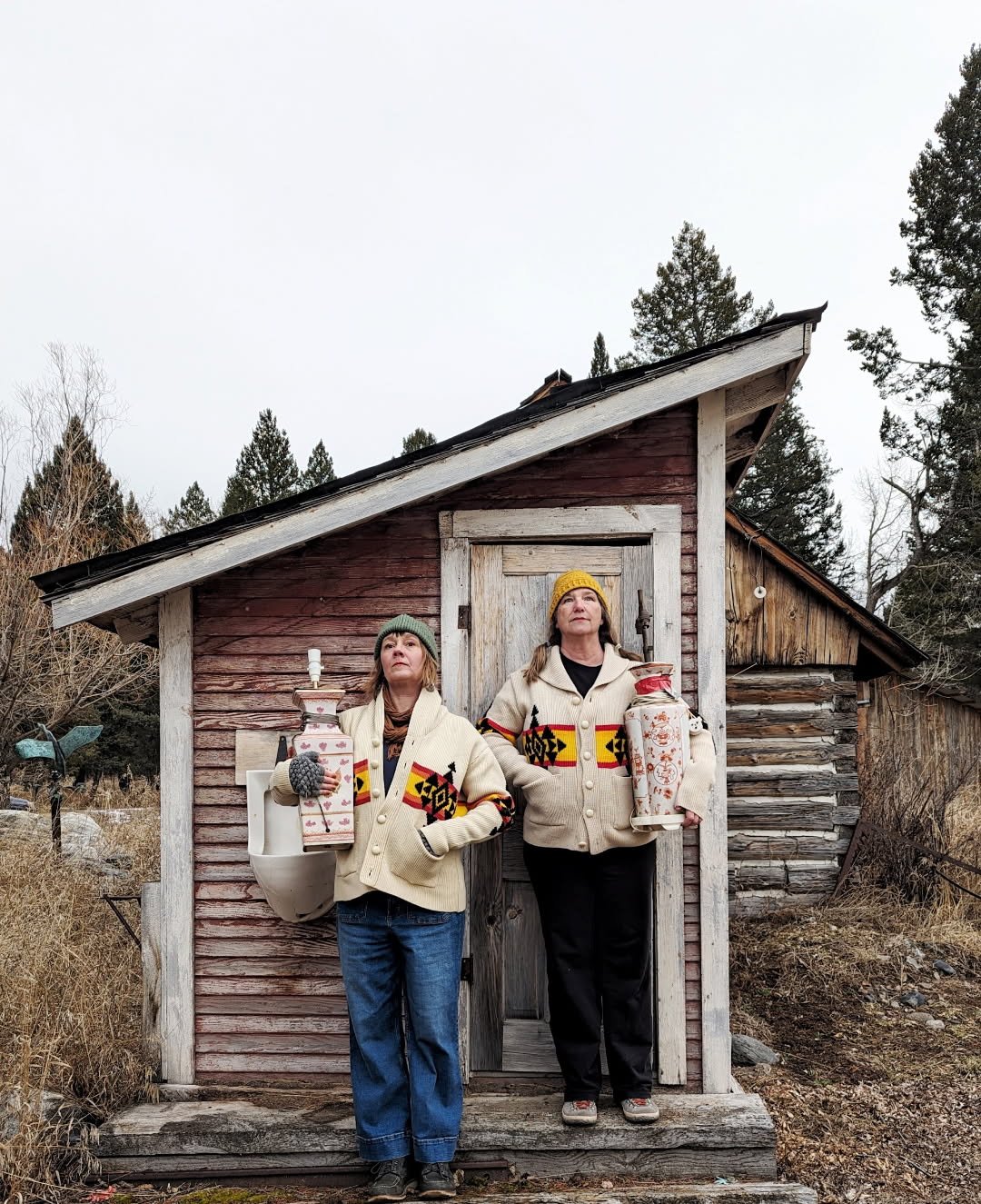 Two women wearing similar patterned sweaters and winter hats, standing in front of a small wooden shed in a rural outdoor setting, holding lanterns.