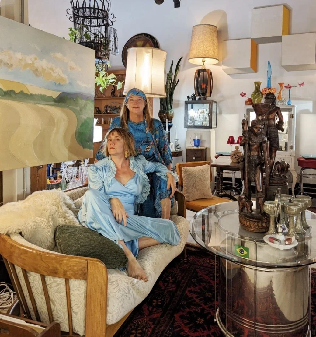 Two women dressed in blue, one sitting on a couch and the other standing behind. They are in a cozy, eclectic living room filled with various decorative items, artwork, and furniture.