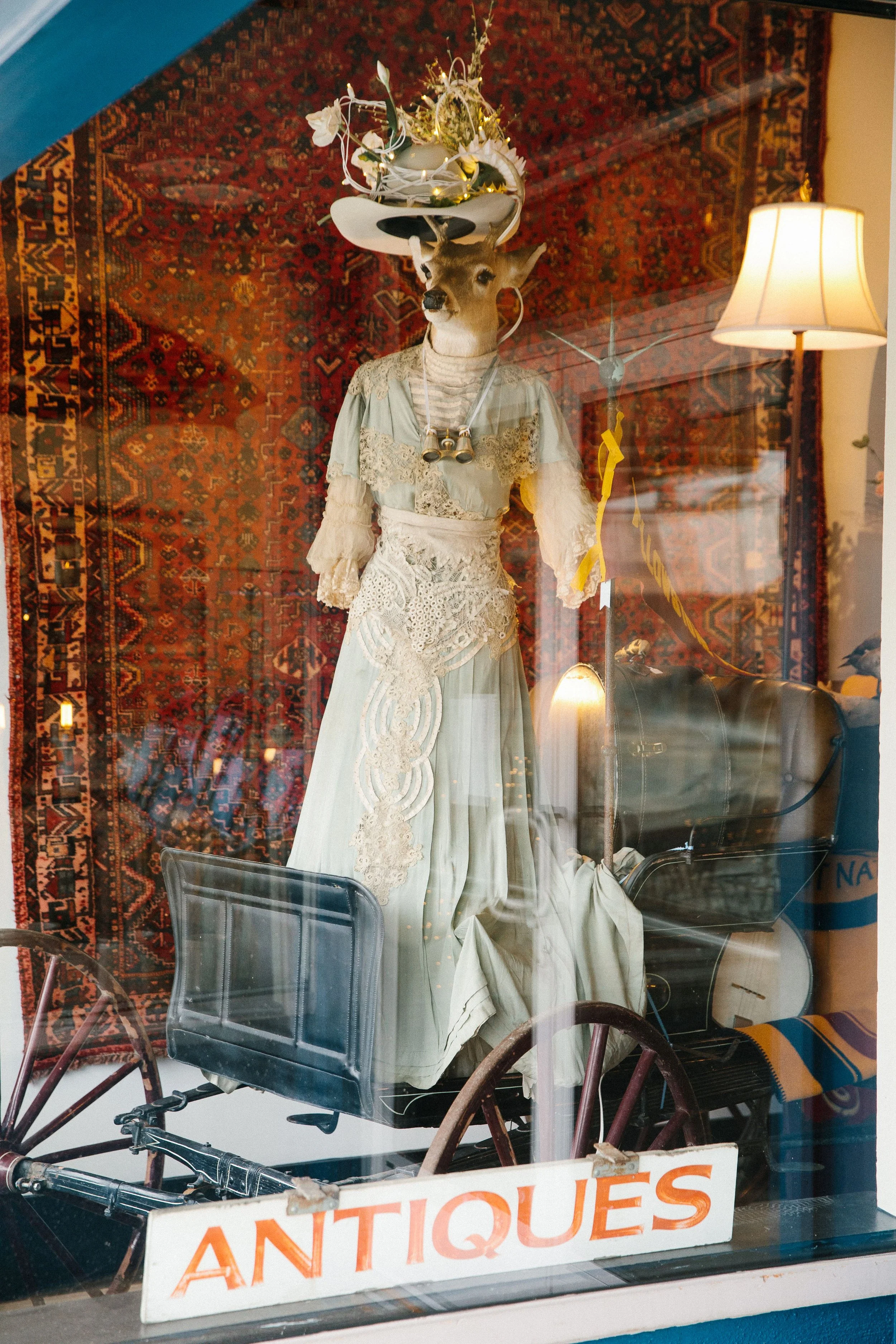 A store display window with an antique mannequin of a deer with a human body dressed in vintage clothing. The mannequin has antlers decorated with string lights and flowers, and is dressed in a lace and satin dress with binoculars around its neck. An