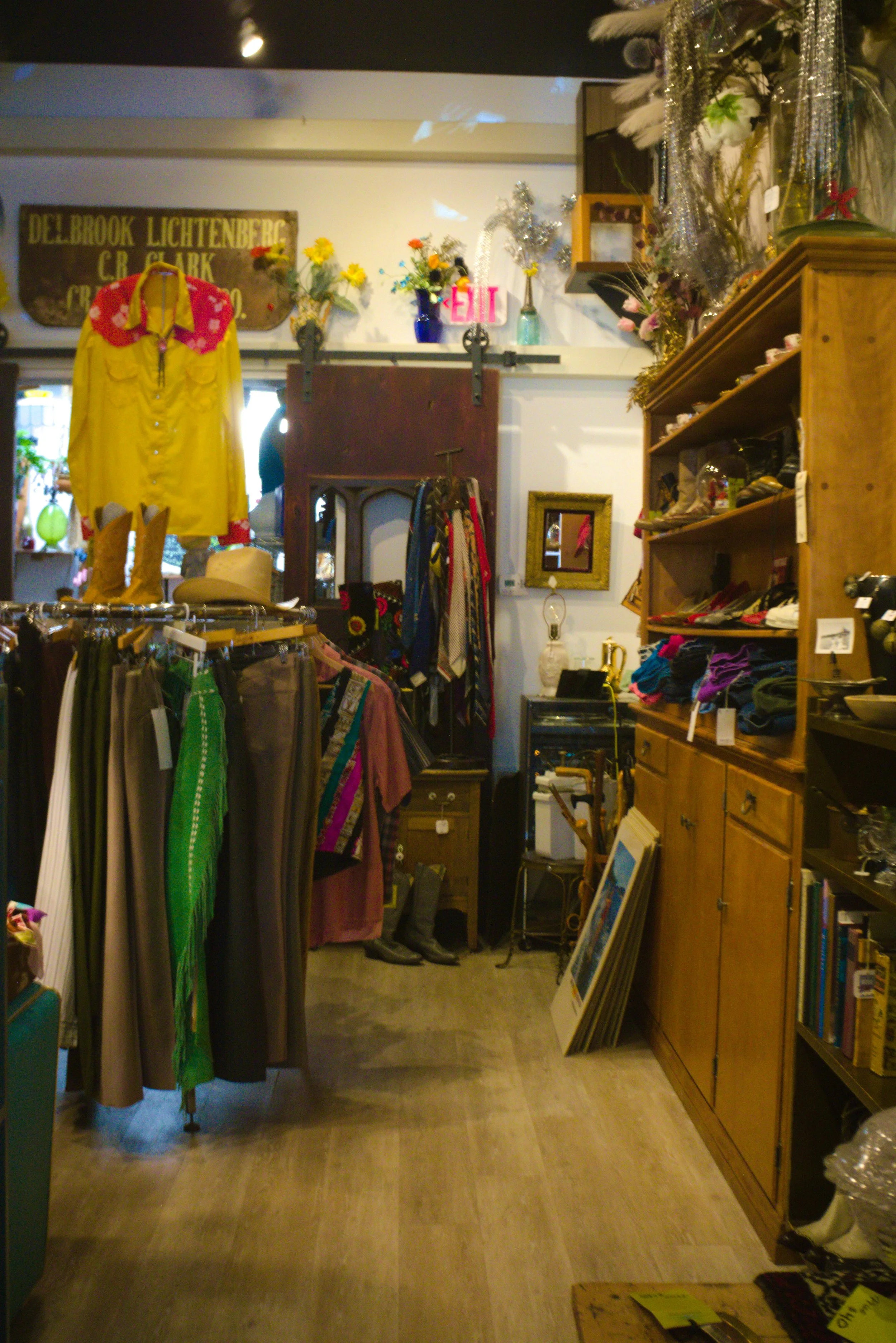 Interior of an antique or thrift shop with clothing, shoes, and decorative items on shelves and racks. A yellow dress with a red collar hangs on a rack, and boots are on the floor. Shelves with books and various decorative objects are on the right si