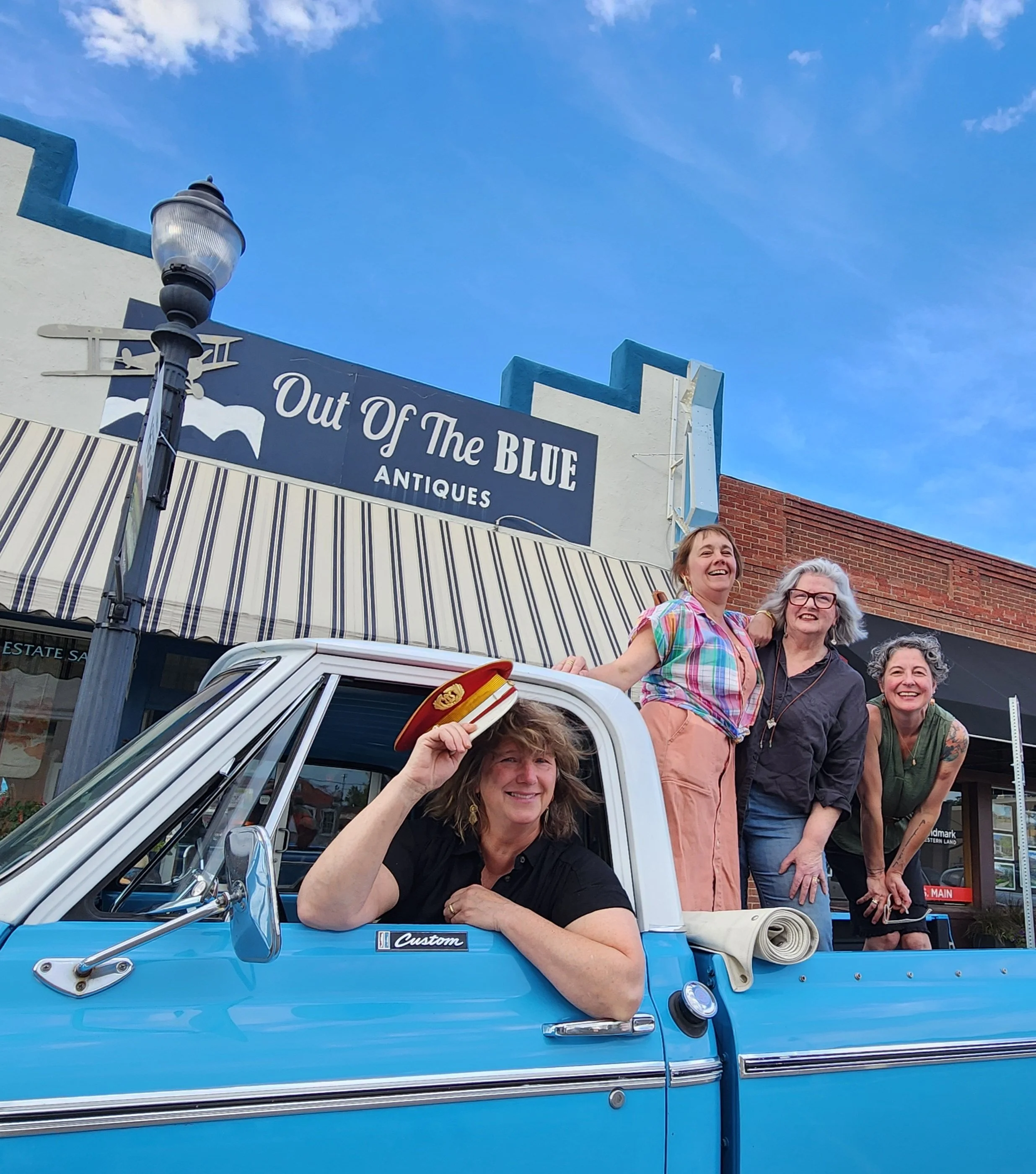 A group of smiling people standing behind a blue vintage truck with a woman holding a red captain's hat in the foreground. In the background, there's a storefront sign for 'Out Of The Blue Antiques' and a clear blue sky with some clouds.