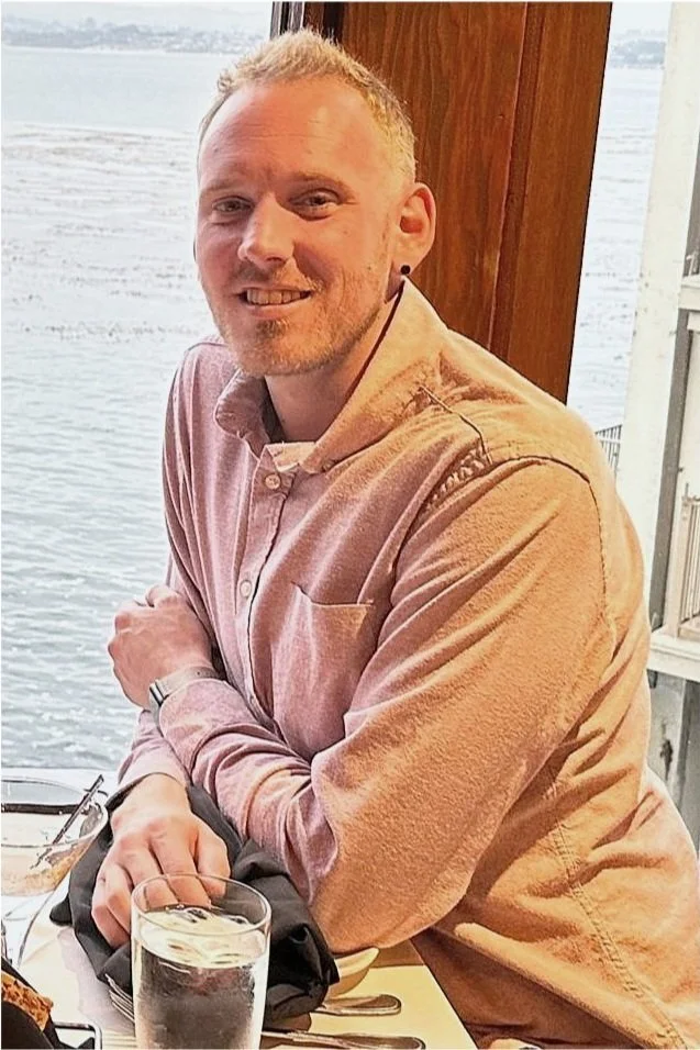 A man with short blonde hair and a beard, smiling, wearing a light pink button-up shirt, sitting at a table near a window with a view of water.