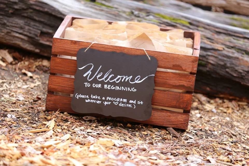 A wooden box with paper inside and a black chalkboard sign reading 'Welcome to our beginning. Please take a program and sit wherever your hearts desires,' placed on the ground among wood chips and foliage.