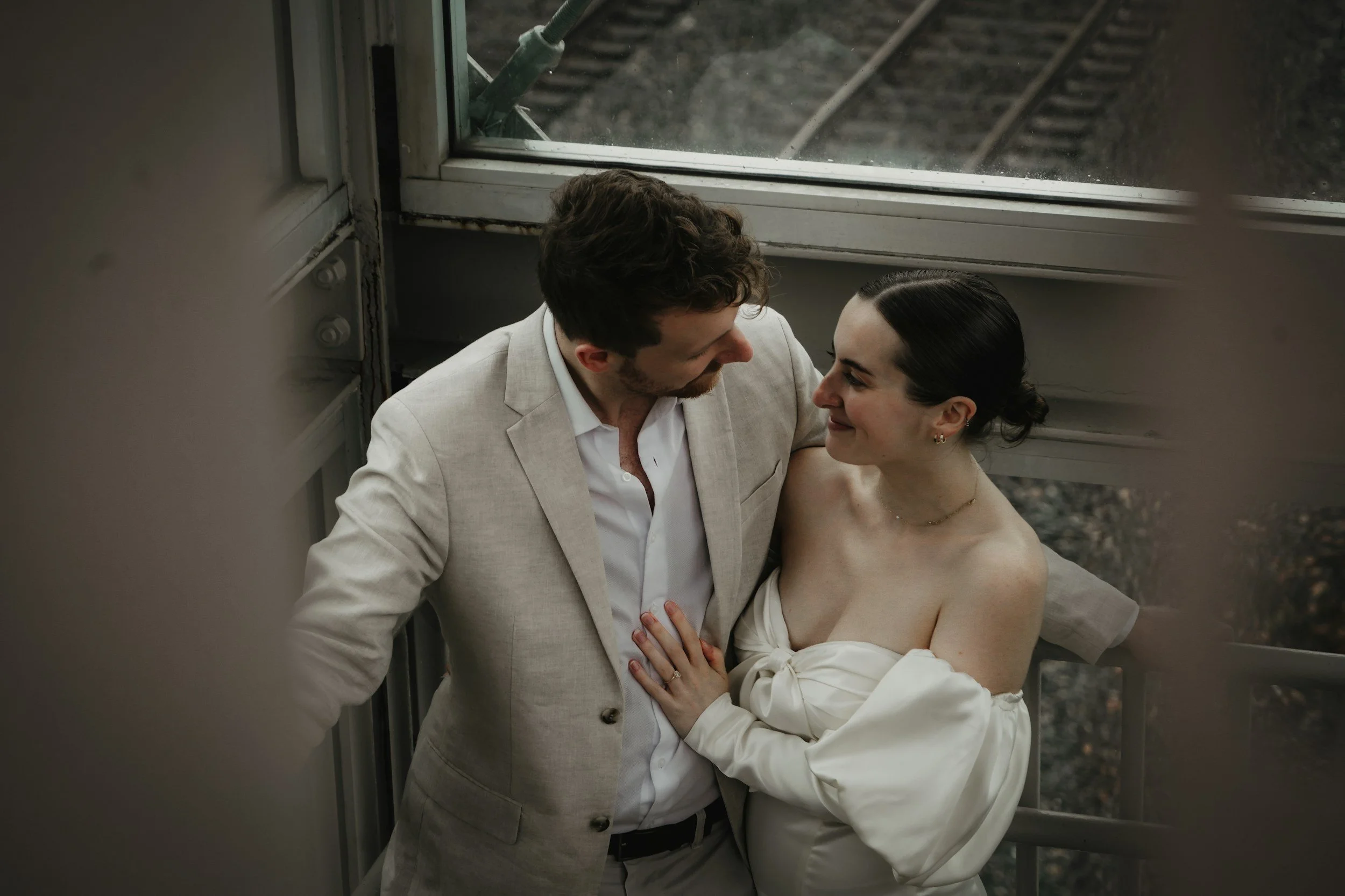 A couple dressed in formal attire sharing an intimate moment, standing on or near train tracks outside, with the man in a light-colored suit and the woman in an off-shoulder white dress.