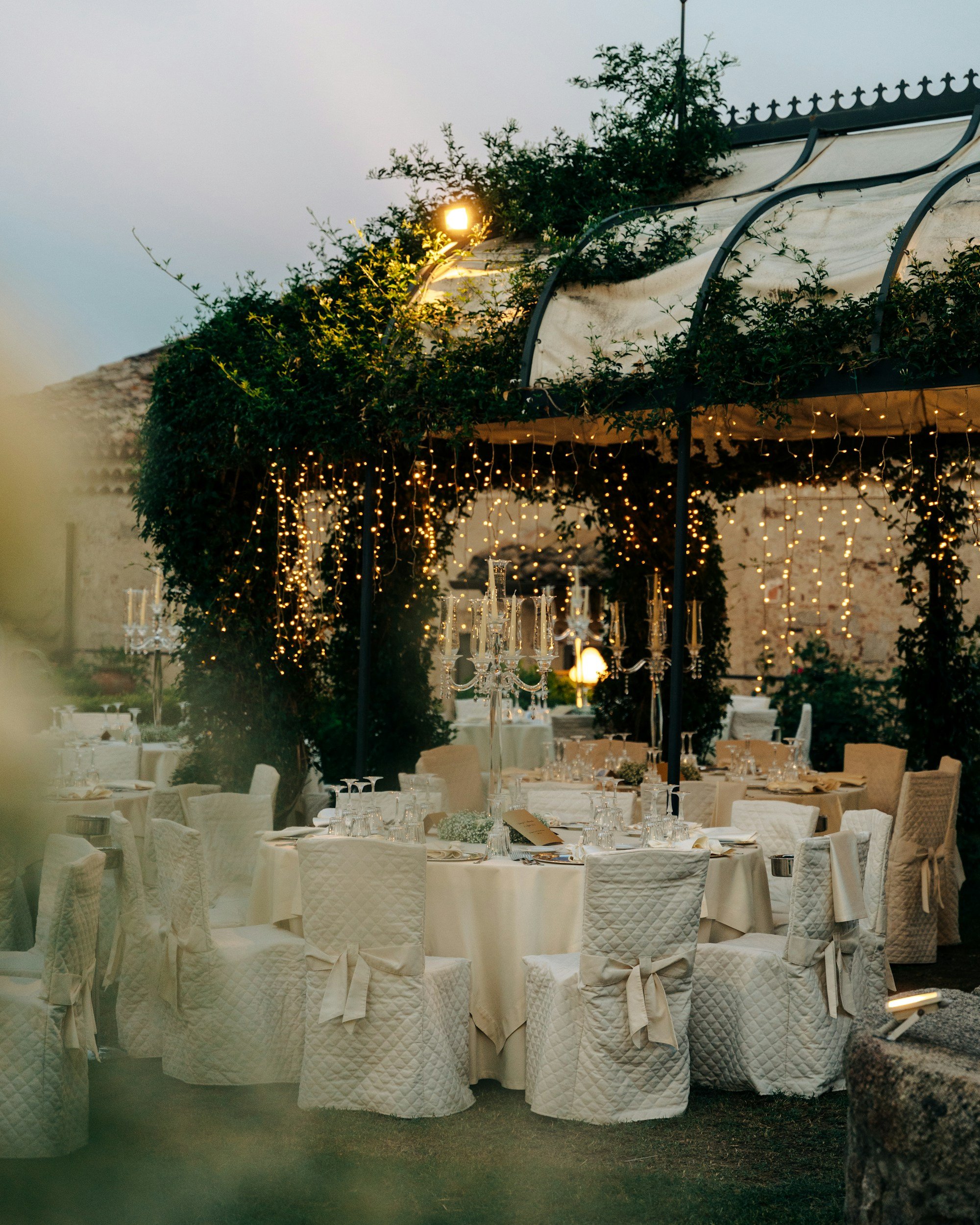 An outdoor event setup with a round table dressed in white tablecloths and chairs covered in white fabric. The area is decorated with string lights hanging from a canopy, and a chandelier centerpiece on the table. In the background, there is a garden or yard area with trees and greenery.
