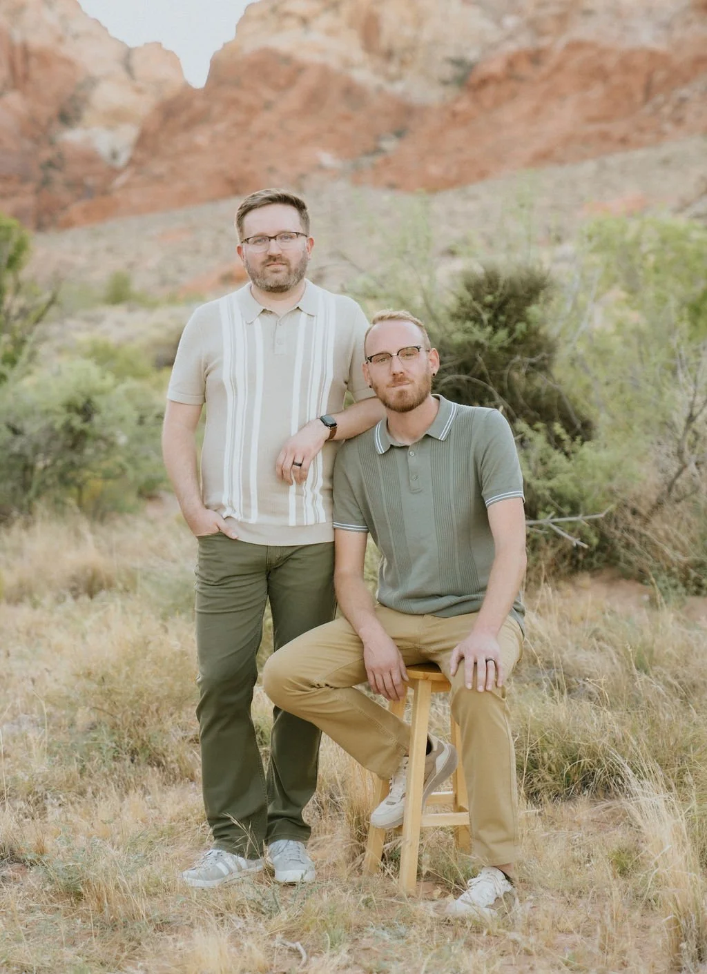 Two men with glasses in a desert landscape with red rock formations and sparse vegetation, one standing and one sitting on a wooden stool.