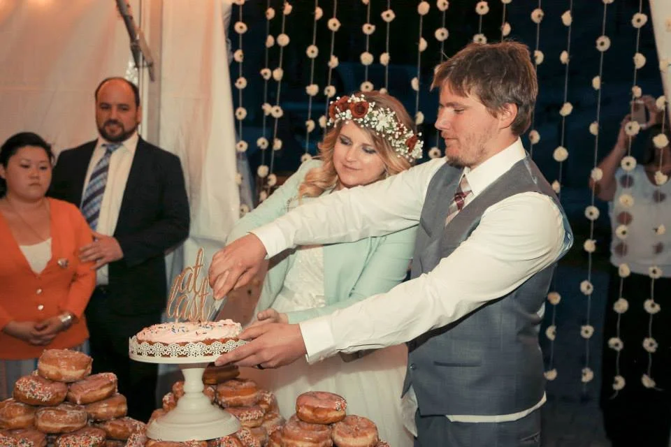 A wedding reception with a couple cutting a wedding cake, surrounded by friends and family, with decorative string lights in the background.