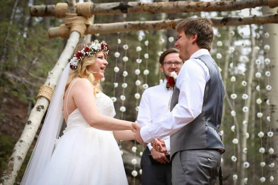 A bride and groom holding hands during their outdoor wedding ceremony, smiling at each other, with an officiant in the background and decorative hanging flowers behind them.