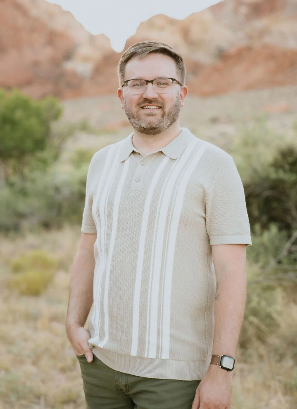 A man with glasses and a beard smiling outdoors, wearing a beige and white striped polo shirt and a smartwatch, with a desert landscape and red rock formations in the background.