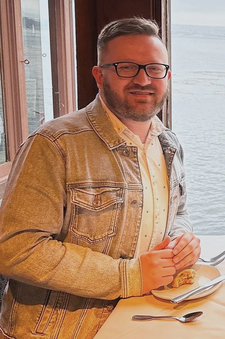 A man with glasses and a beard, wearing a denim jacket and a light yellow shirt, sitting at a table near a window with a view of the water, holding a dessert on a plate.