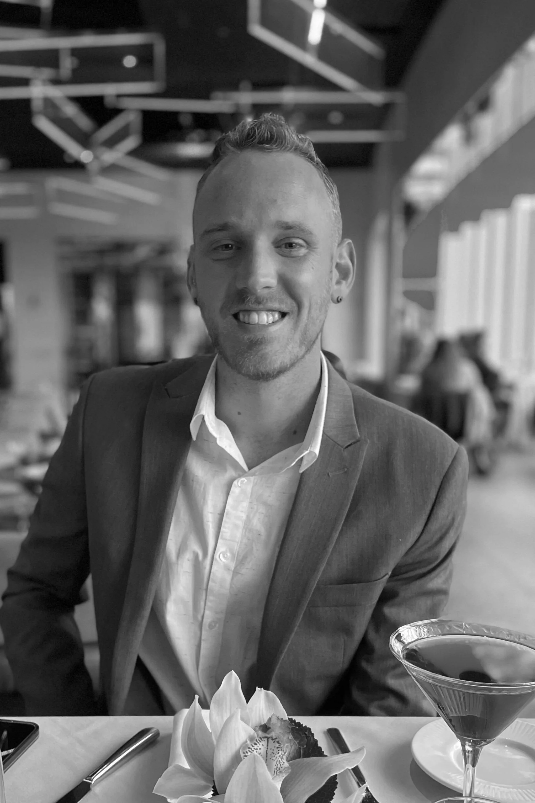 A man in a suit smiling at a dining table in a restaurant, with a cocktail glass, a flower, a plate of food, and utensils visible in the foreground, and modern decor in the background.