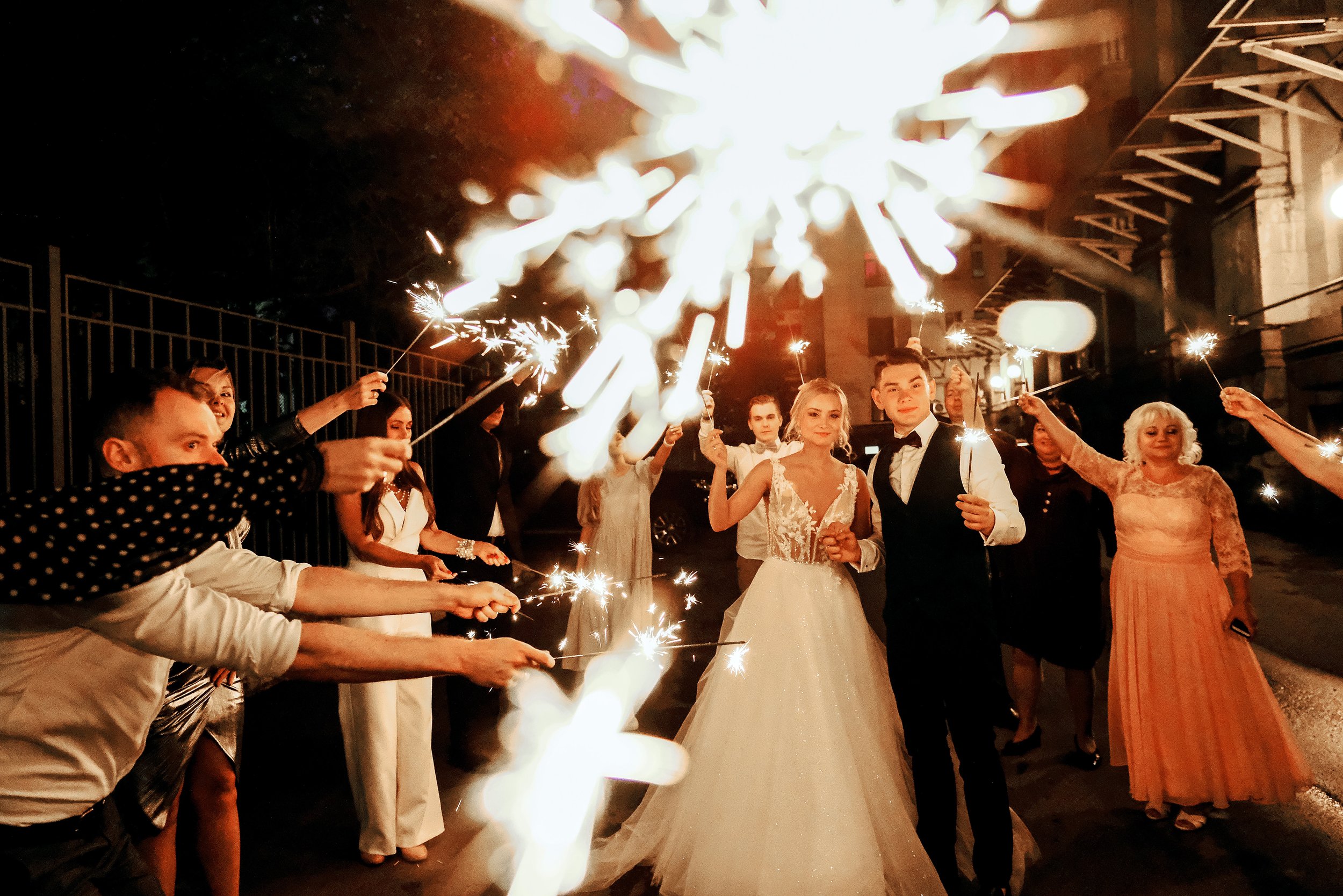 People celebrating at night with sparklers. The bride and groom are in the center, surrounded by friends and family, with fireworks and festive lights in the background.