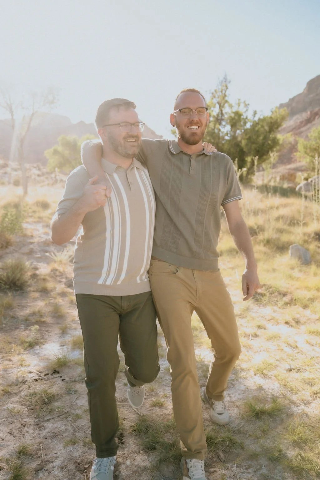 Two men smiling and walking outdoors on a sunny day in a desert landscape with rocks and bushes, with one man having an arm around the other's shoulder.