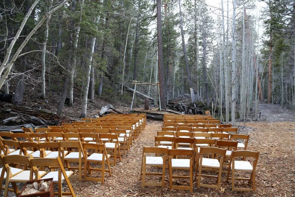 Wooden chairs arranged in rows on a forest floor, with a wooden arch and string lights in the background, set up for an outdoor event in a wooded area.