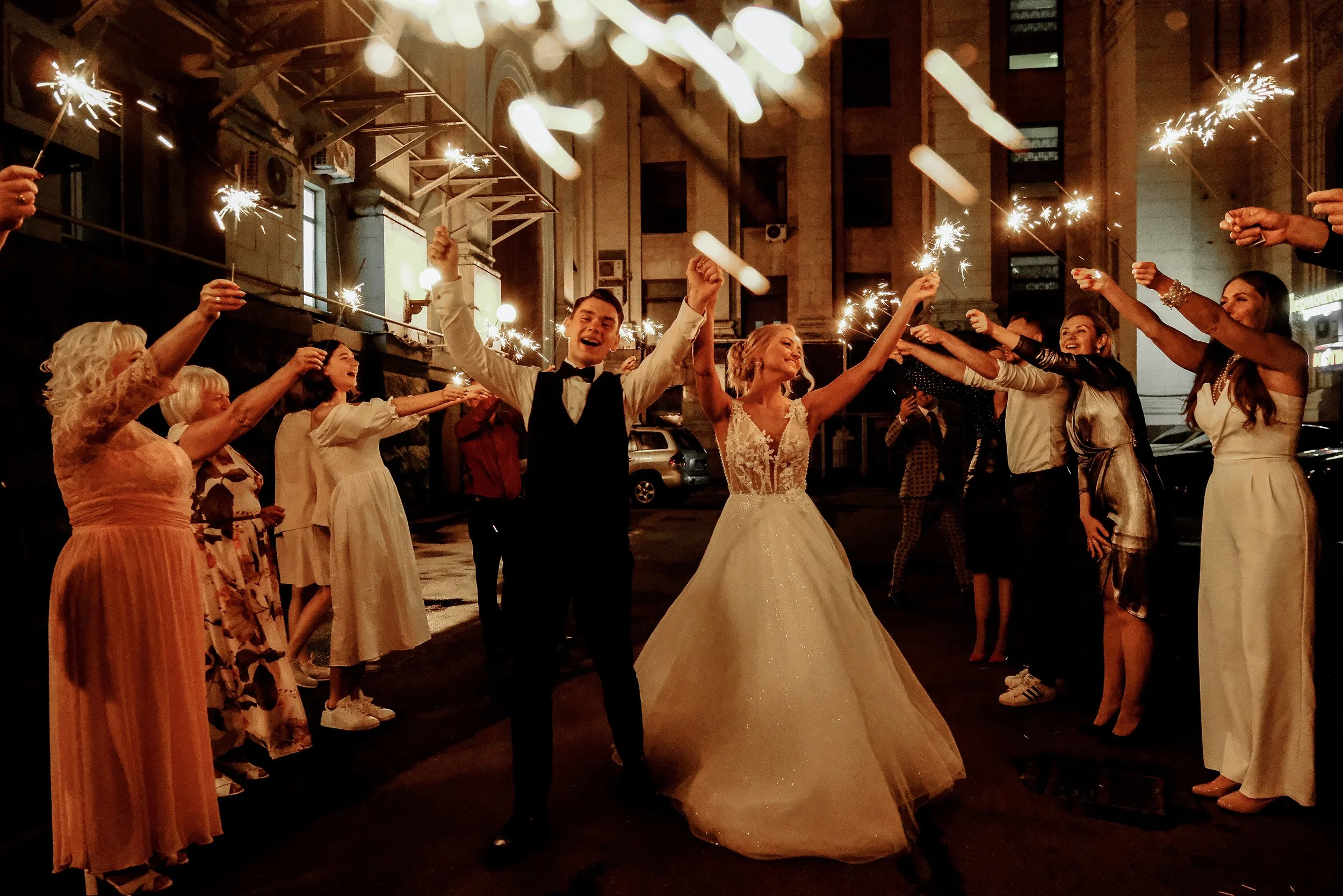 People celebrating at night, some holding sparklers and candles, with a bride and groom in the center. The bride wears a white wedding dress, and the groom wears a tuxedo, smiling joyfully. The scene appears festive, possibly at a wedding reception or celebration, in an urban setting with buildings in the background.