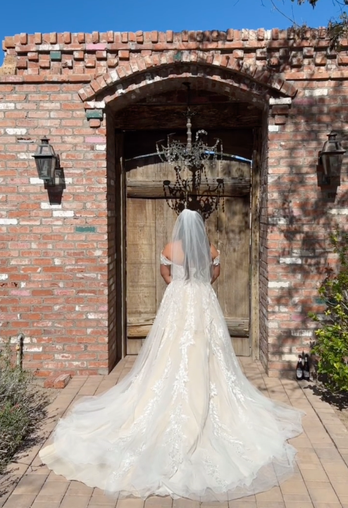 Bridal figure in white wedding dress with long train and veil, facing a wooden door set in a brick wall, with outdoor lanterns on each side and a chandelier hanging above.