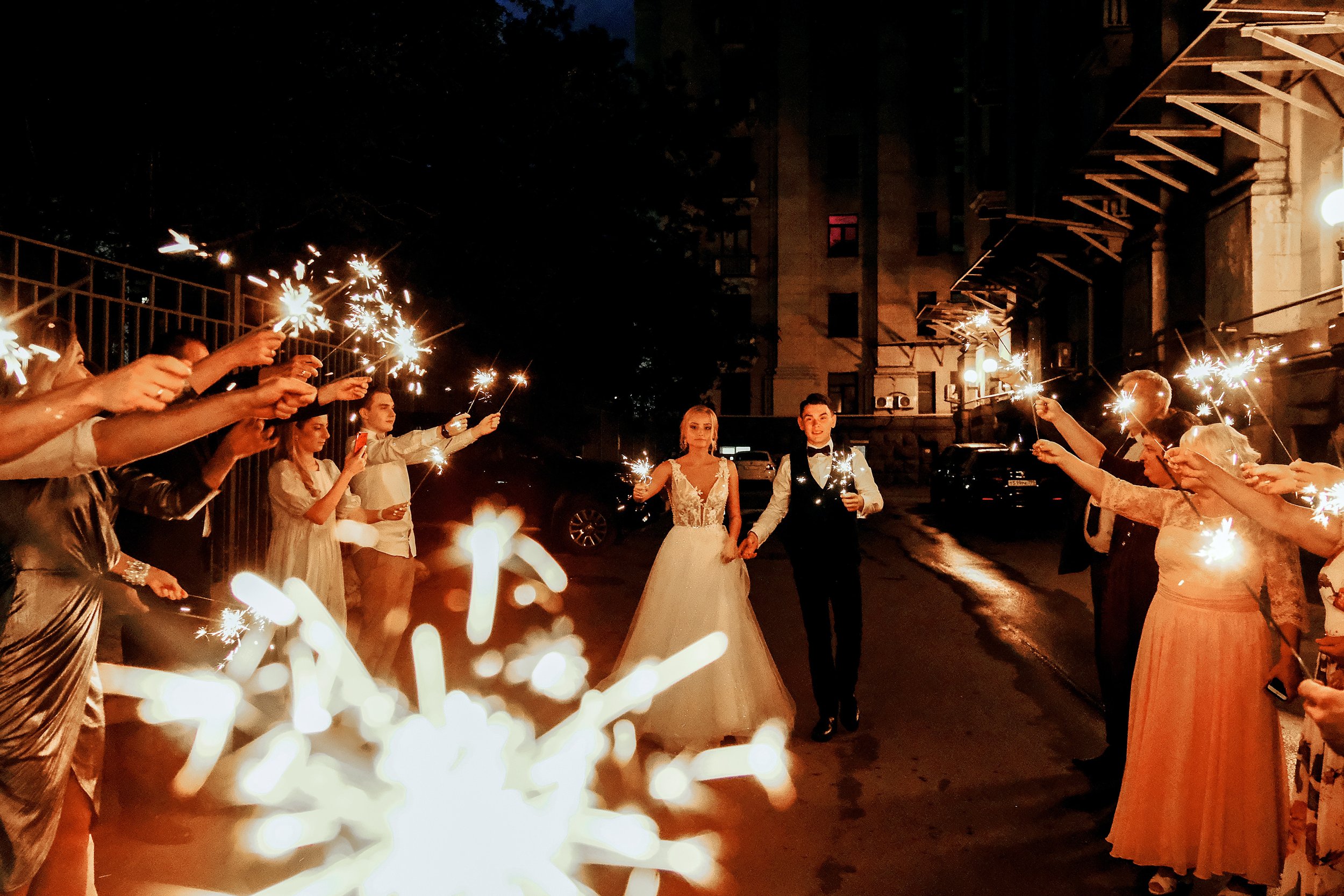A bride and groom standing together during a nighttime celebration, surrounded by guests holding sparklers. The scene appears festive and romantic outside a building.