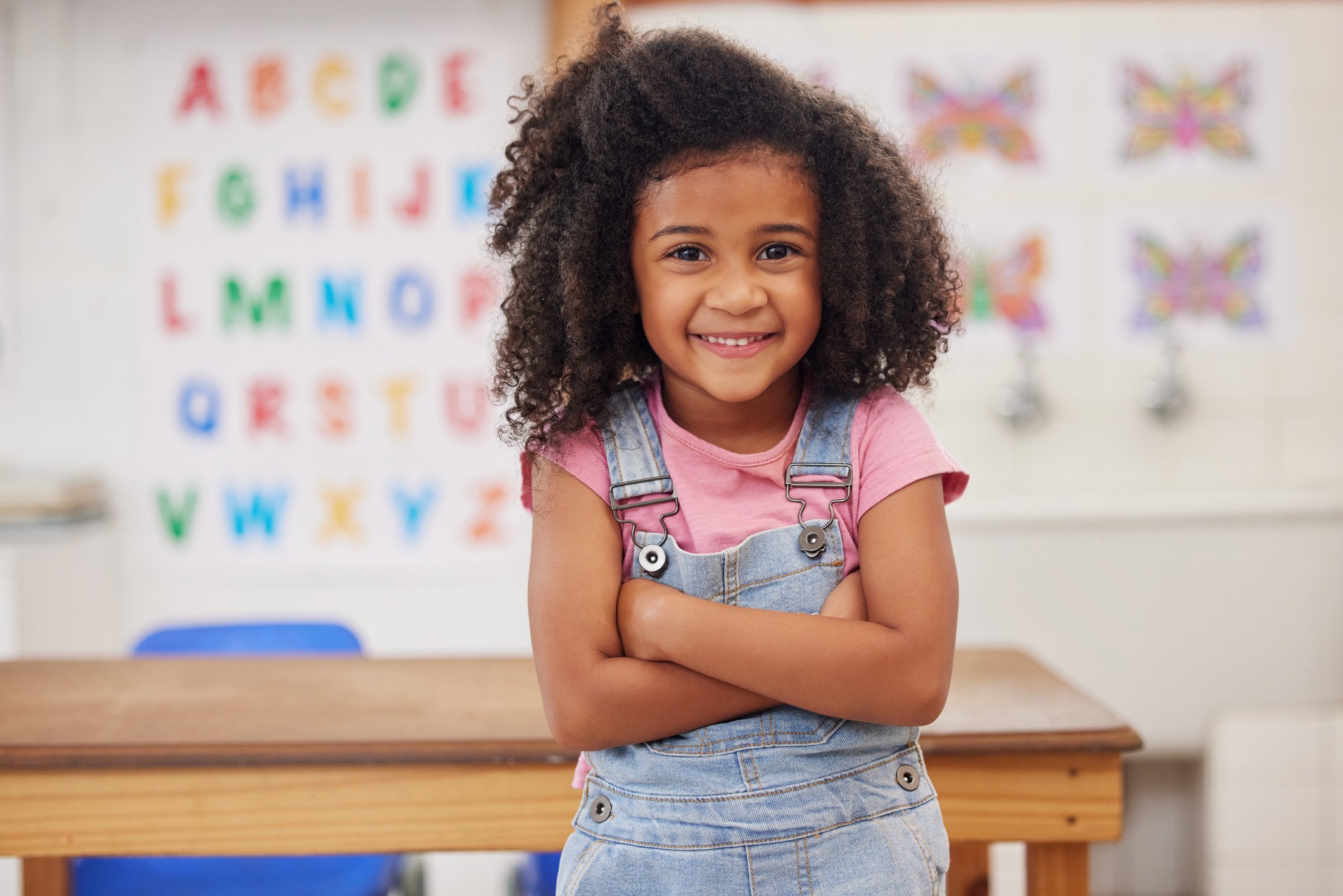 Young school girl is smiling in a classroom with alphabet posters and butterfly artwork in the background.