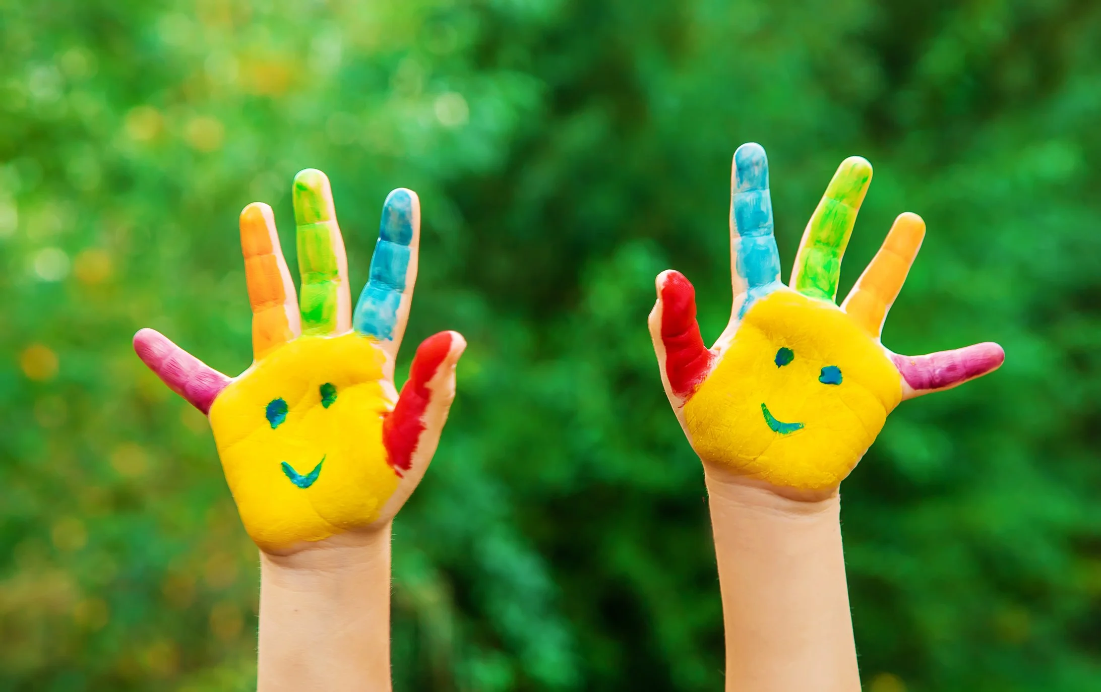Two hands painted with bright color held up against a green outdoor background.