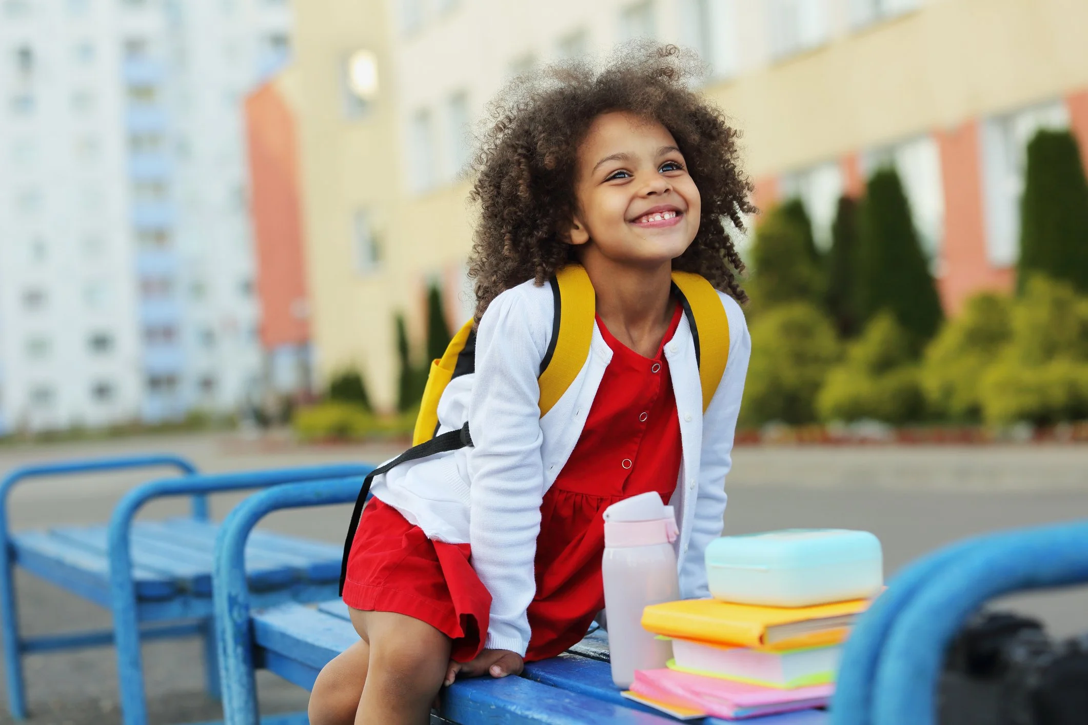 A smiling young girl with is sitting on a bench with her backpack and lunch outside of school.