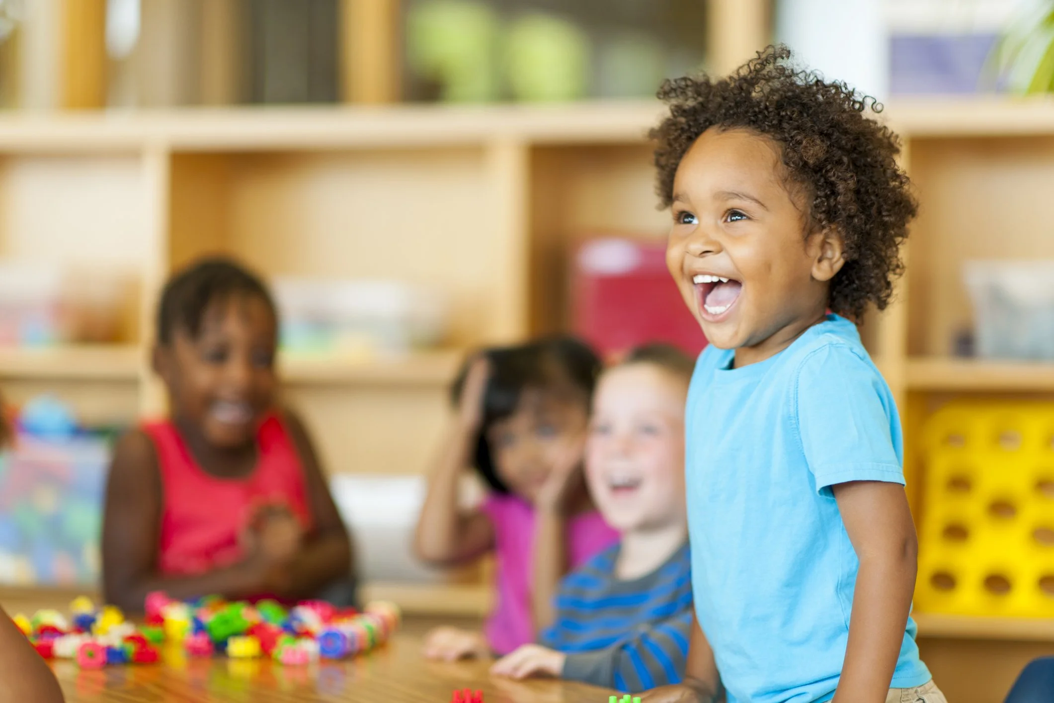 A young boy is laughing and smiling in a classroom with kids blurred in the background.