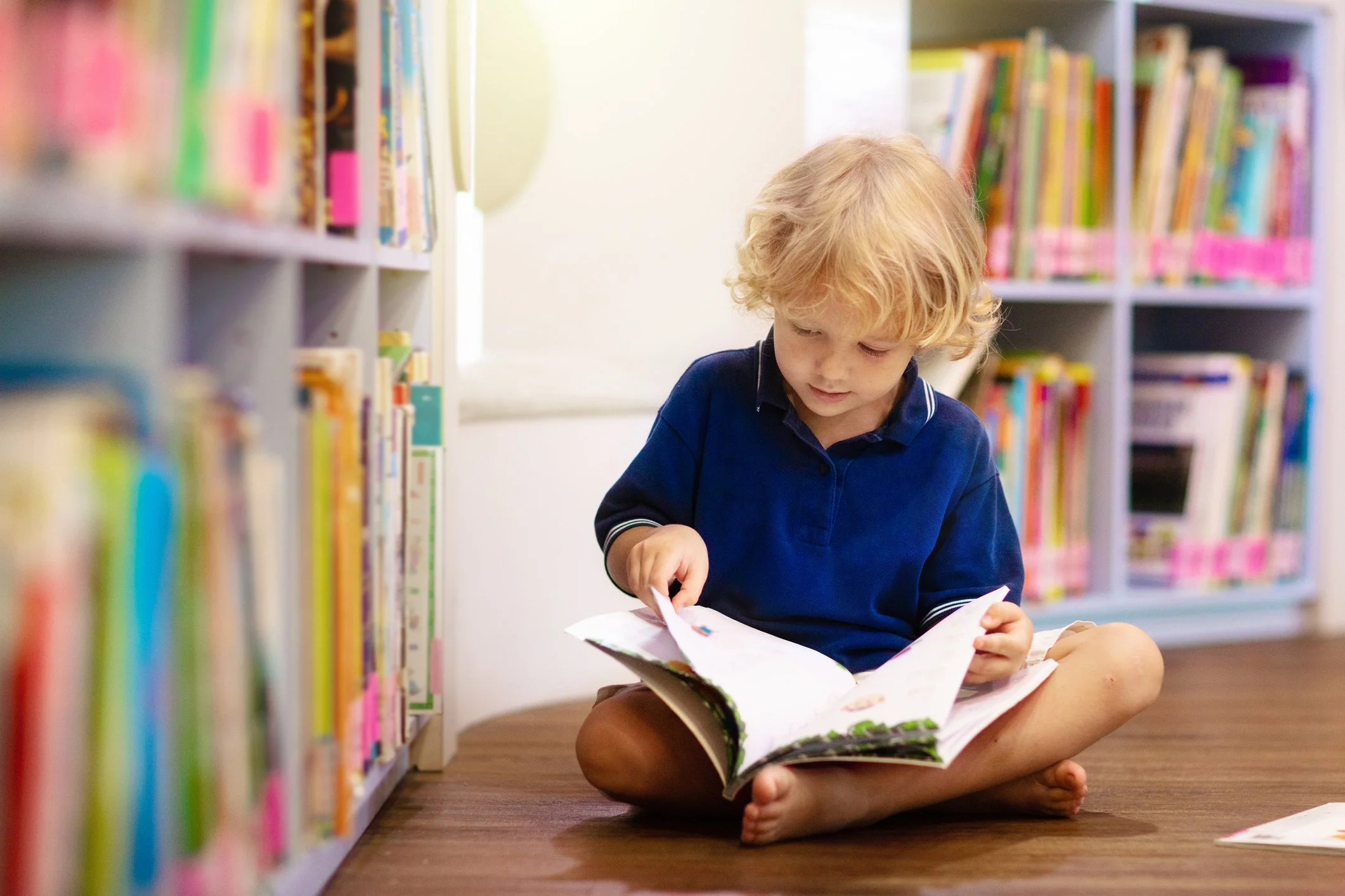 A young boy is sitting on the floor of a library reading a book.