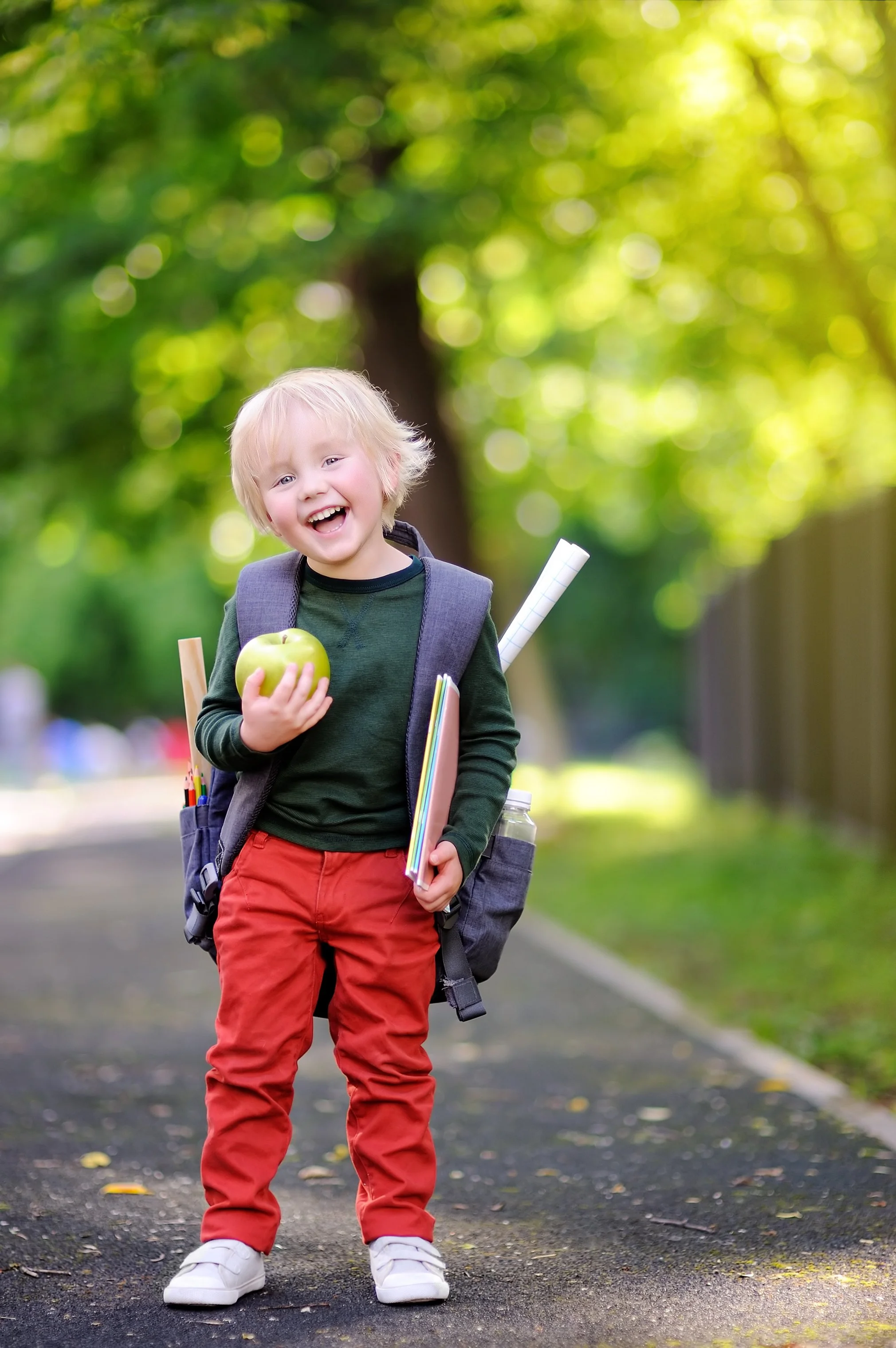 A preschool boy is holding school supplies and wearing a school backpack outside.