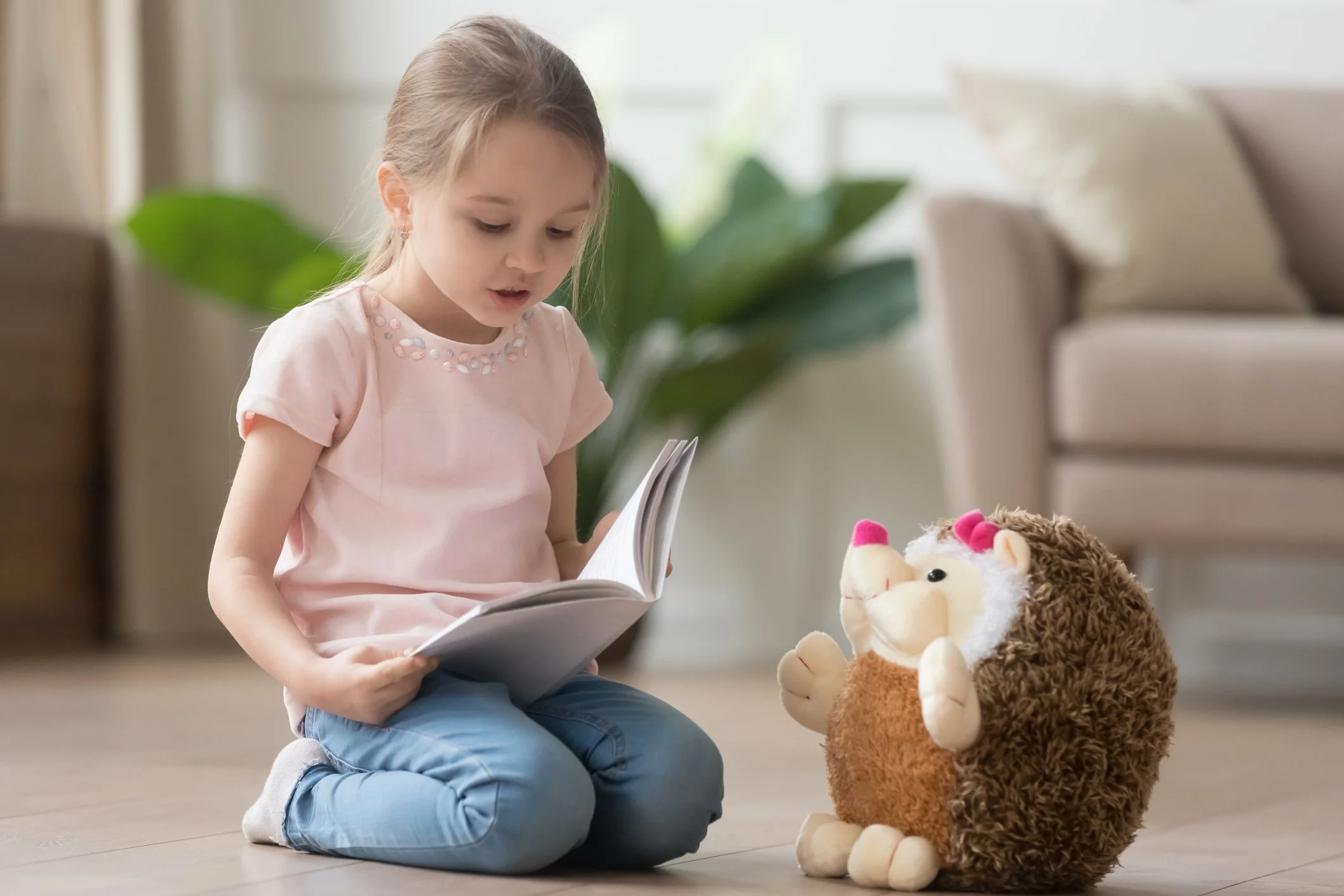 A preschool girl sitting on the floor reading a book to a hedgehog stuffed animal.