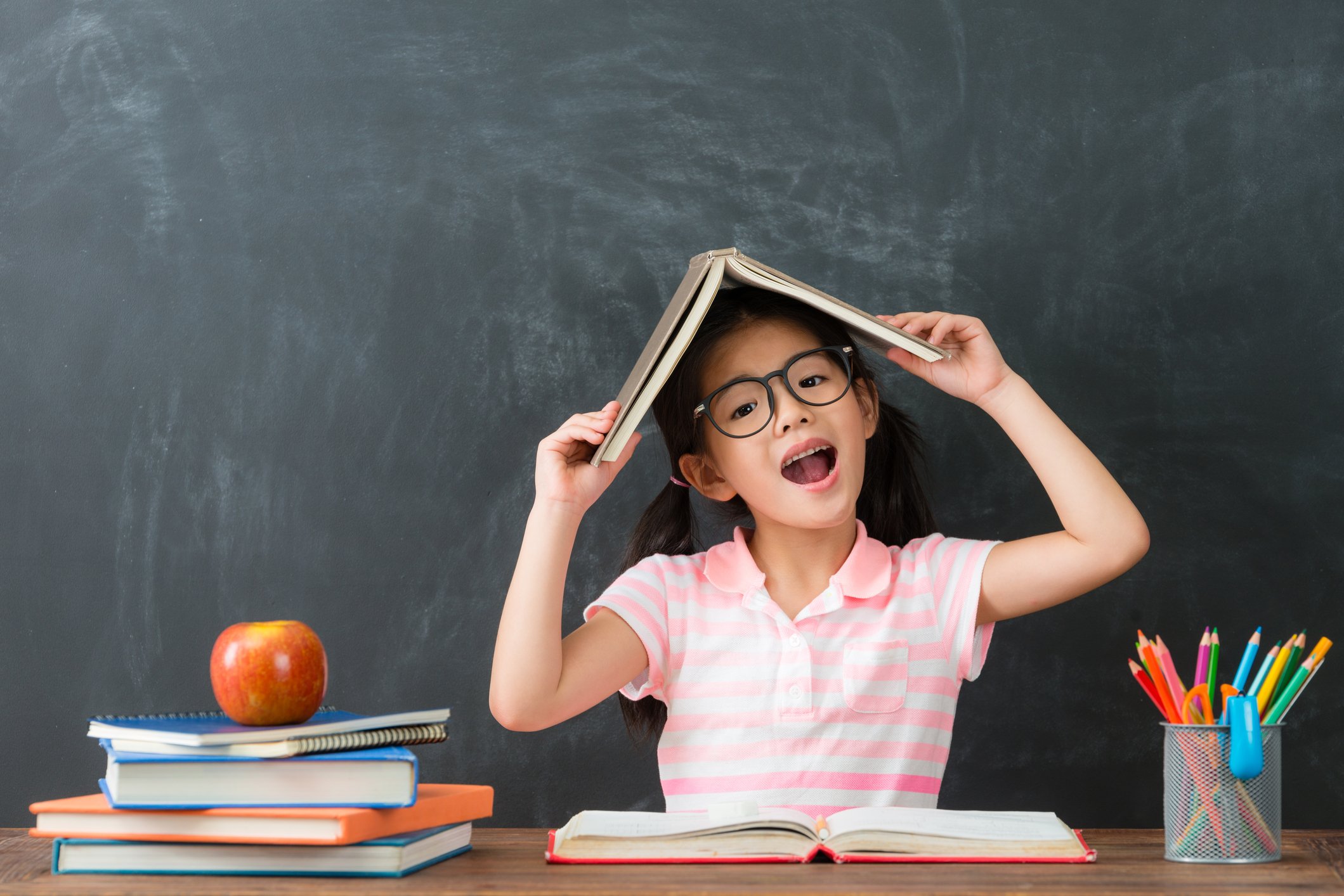 A young girl holding a book over her head while sitting at a desk with school supplies.
