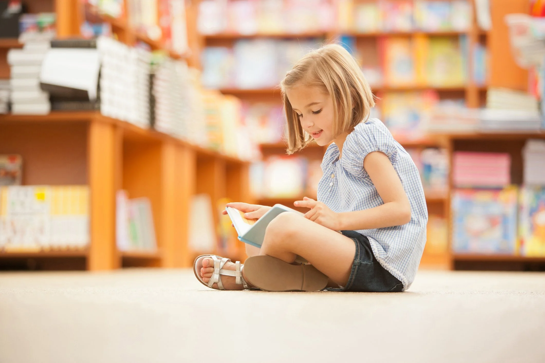 A young girl sitting on the floor of a library reading a book.