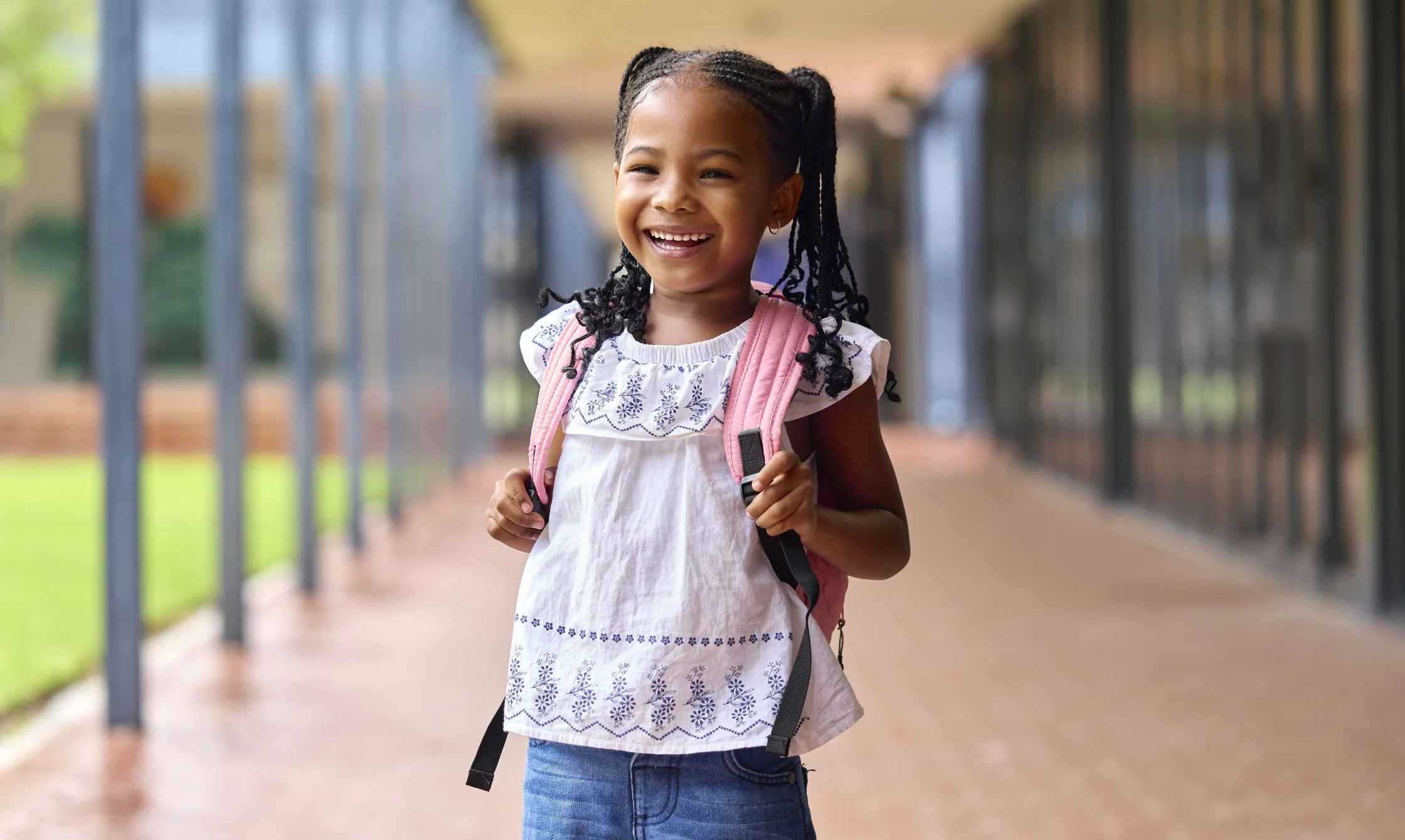 A smiling young girl is standing outside of school carrying a pink backpack.