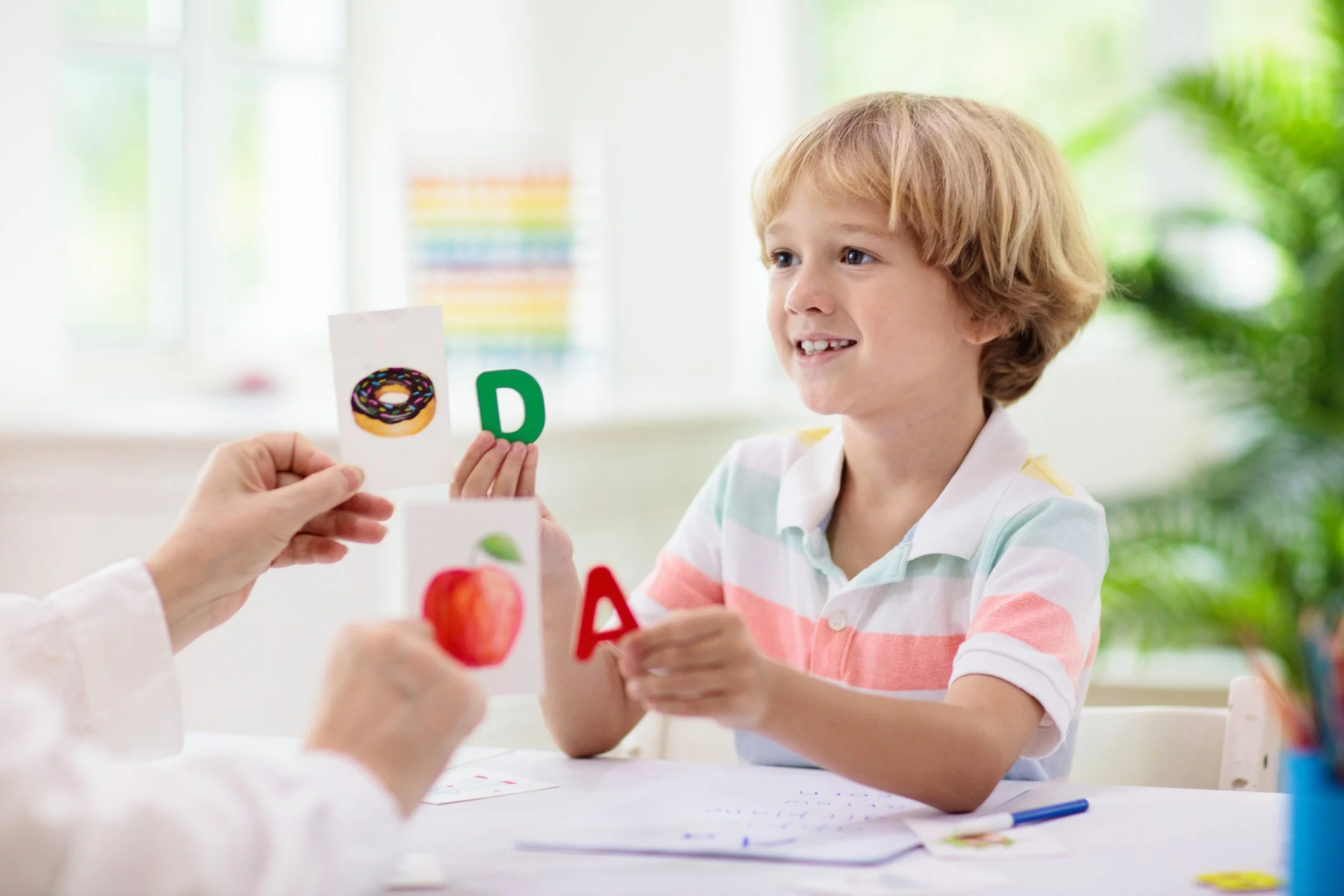 A young boy is holding up alphabet and picture cards at a desk.