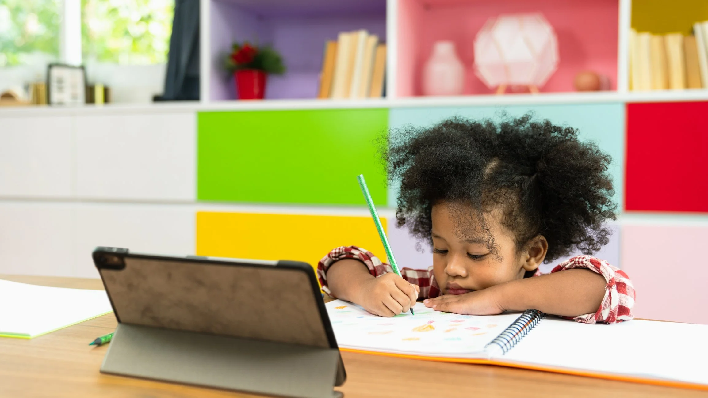 A young girl is sitting at a desk and writing on paper with an ipad in front of her.