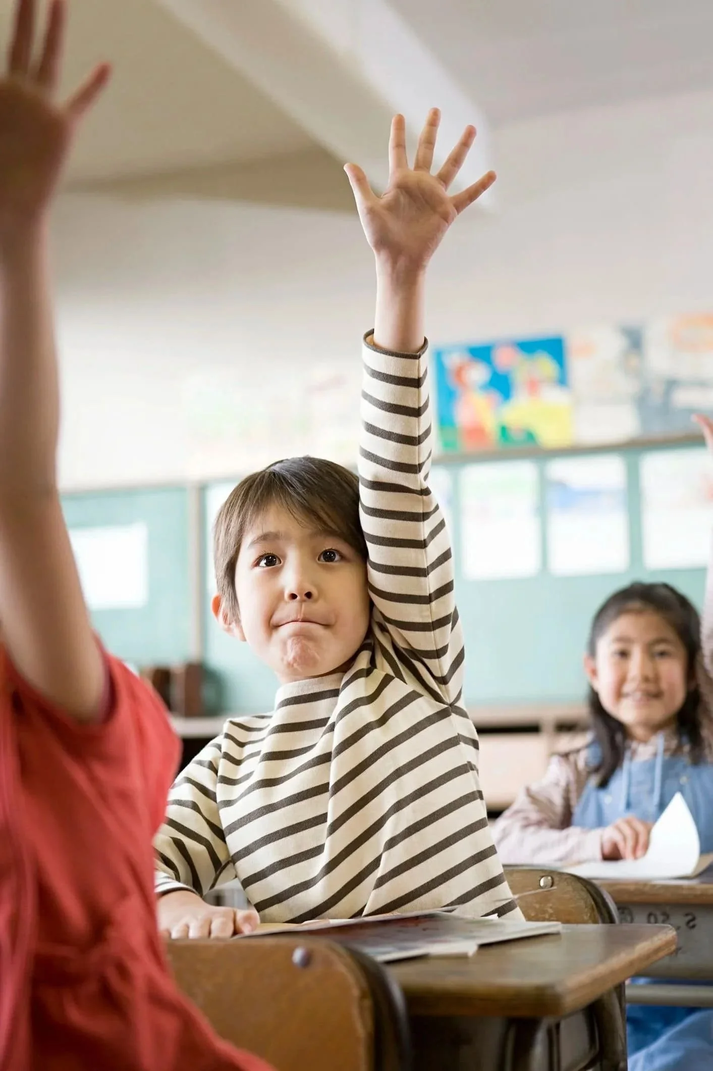 An elementary boy is sitting at a desk raising his hand in a classroom of kids.