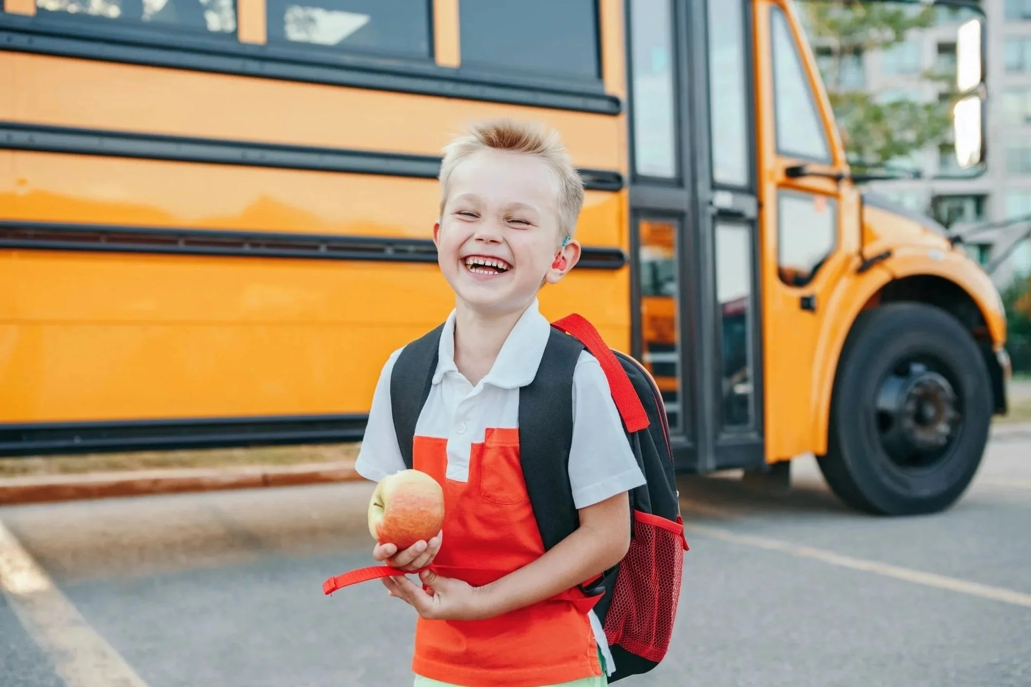 Young boy wearing a backpack is smiling while standing in front of a school bus.