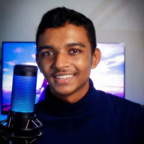 A young man smiling in front of a microphone with a computer monitor in the background.