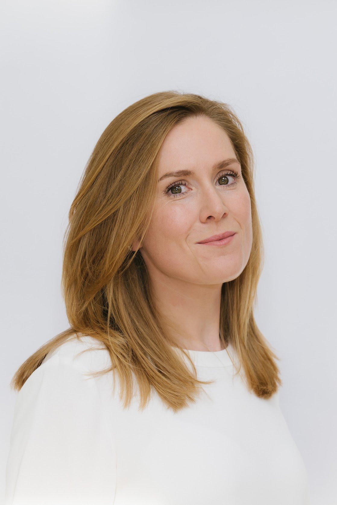 Portrait of a woman with light skin and red hair, wearing a white top, smiling softly against a plain white background.