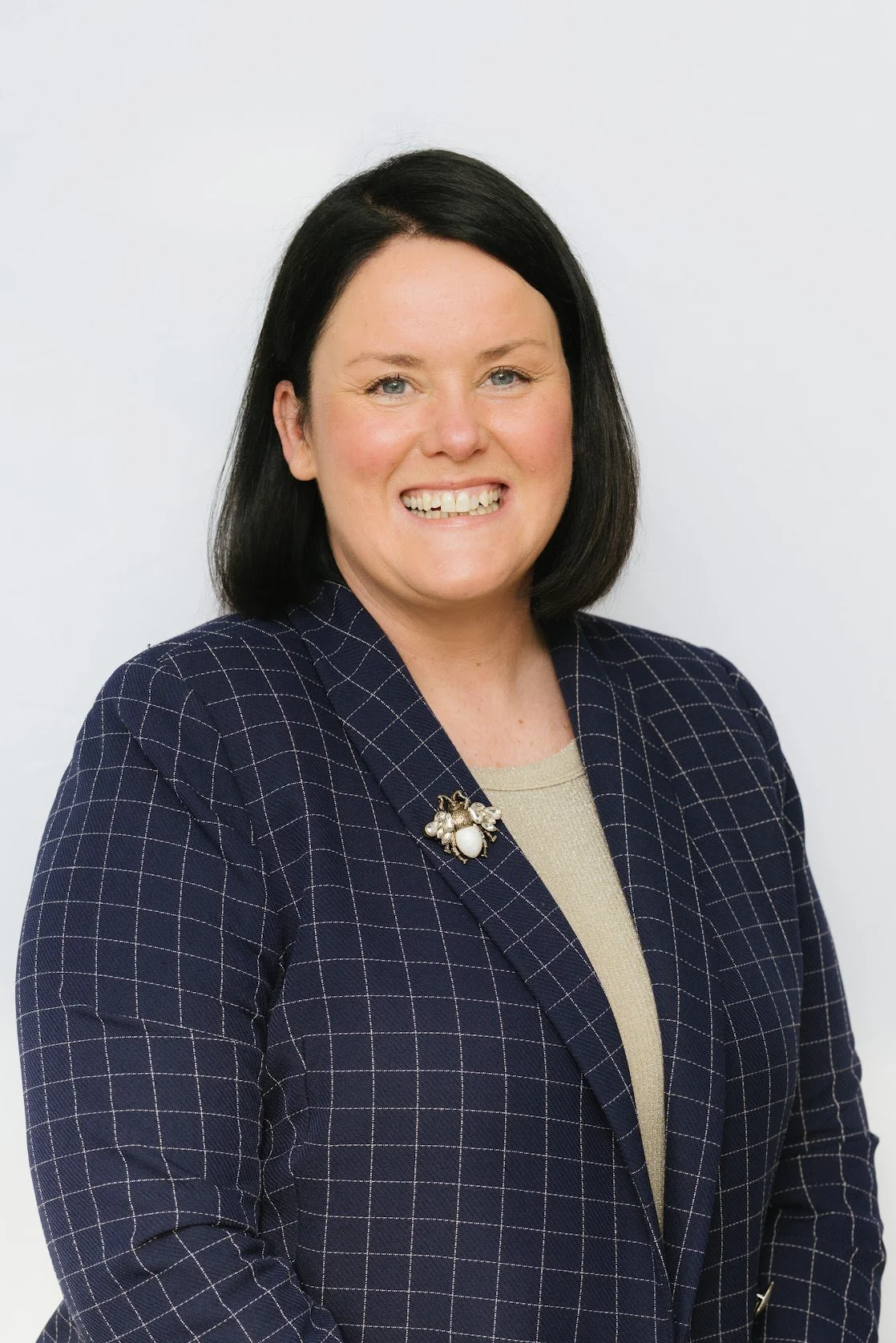 Professional portrait of a smiling woman with dark hair, wearing a navy checkered blazer and decorative brooch, against a plain white background.