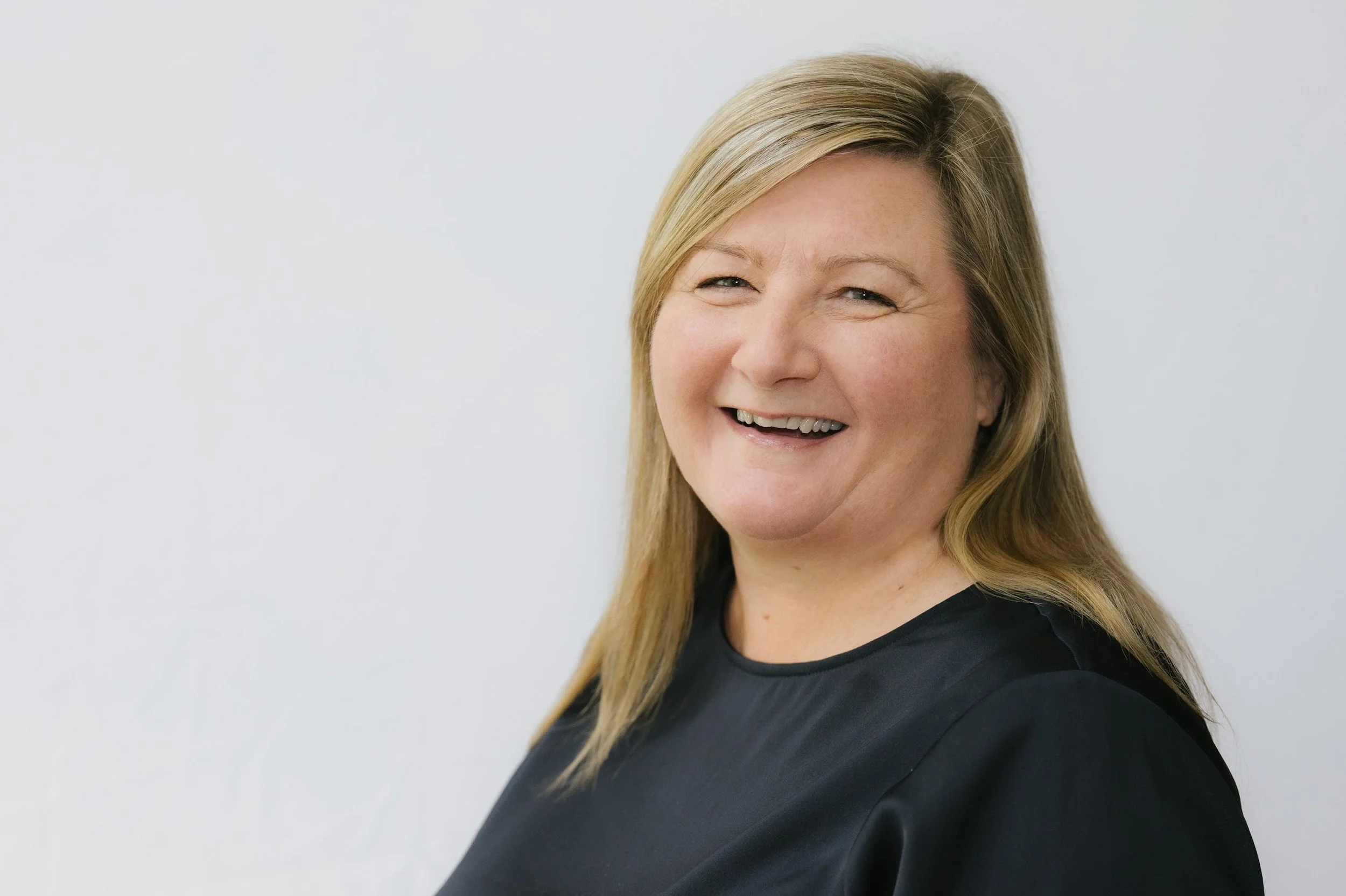A woman with blonde hair smiling and wearing a black top, posing against a plain white background.